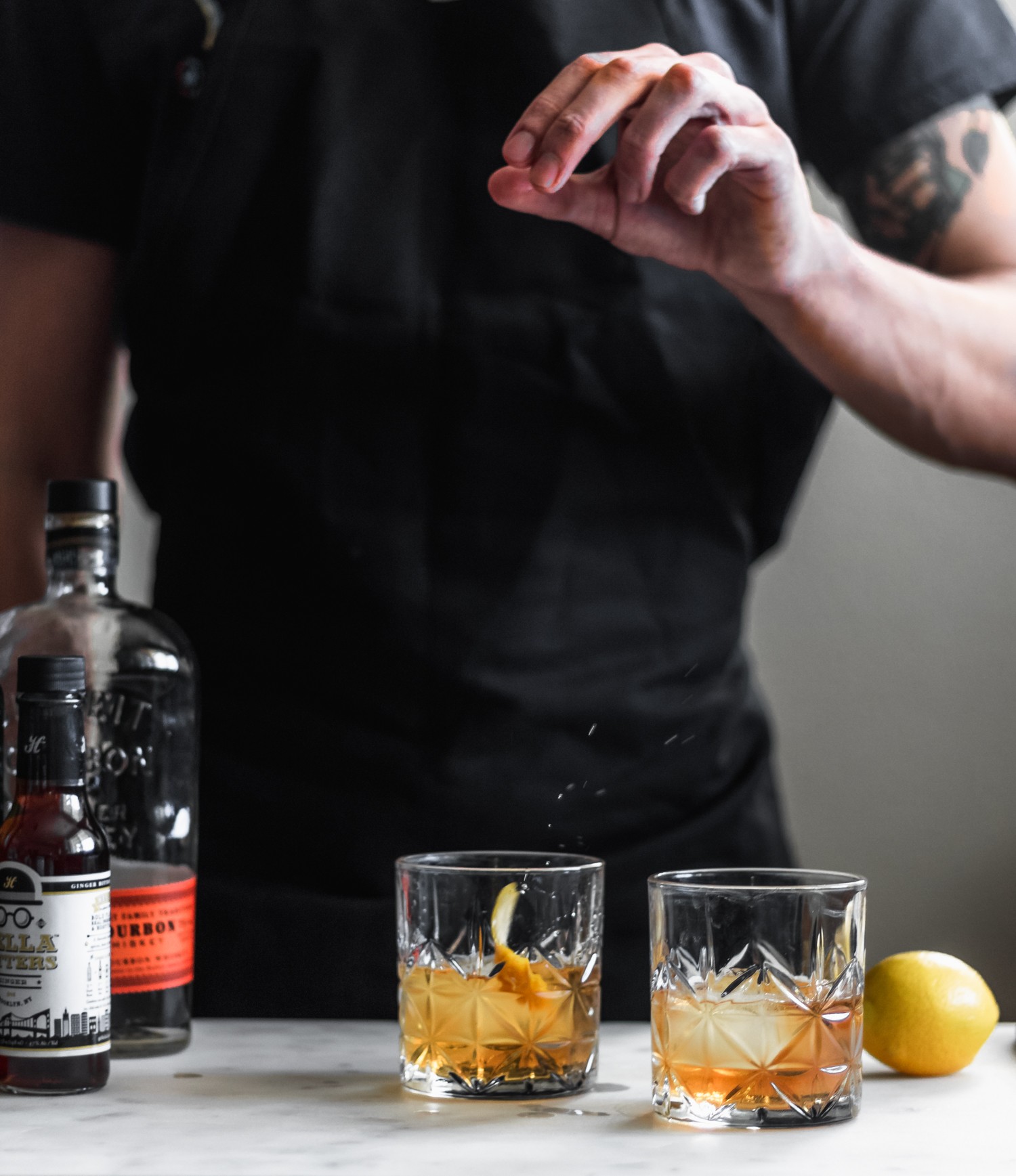 A side image of a man wearing a black apron dropping a lemon peel into a whiskey cocktail on a marble counter next to a bottle of whiskey and bottle of bitters.