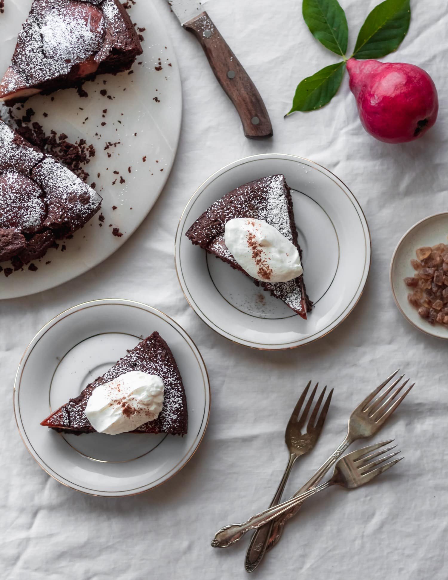 An overhead image of two white plates with chocolate cake slices and whipped cream on a white linen next to the whole cake, vintage forks, and a red pear.