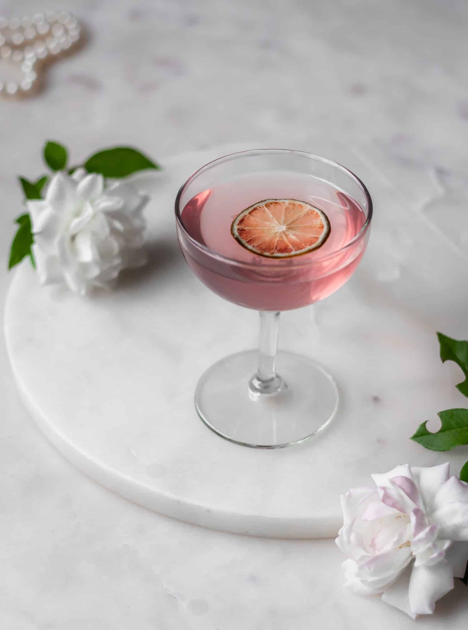 A prickly pear vodka sour in a coupe glass on a marble counter next to white roses and pearls.