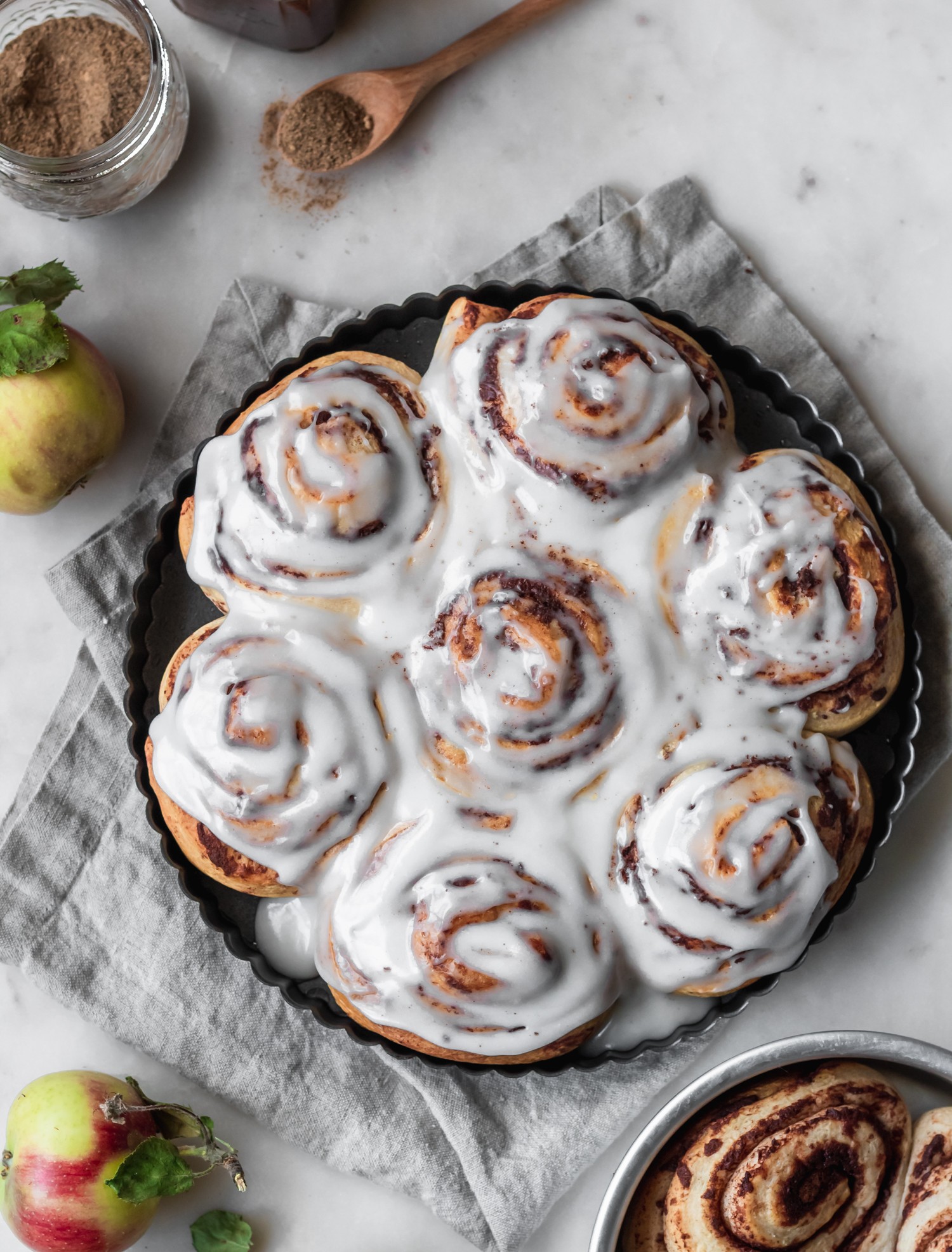 A closeup image of apple butter cinnamon rolls with brown butter icing on a marble counter next to a grey napkin and apples.