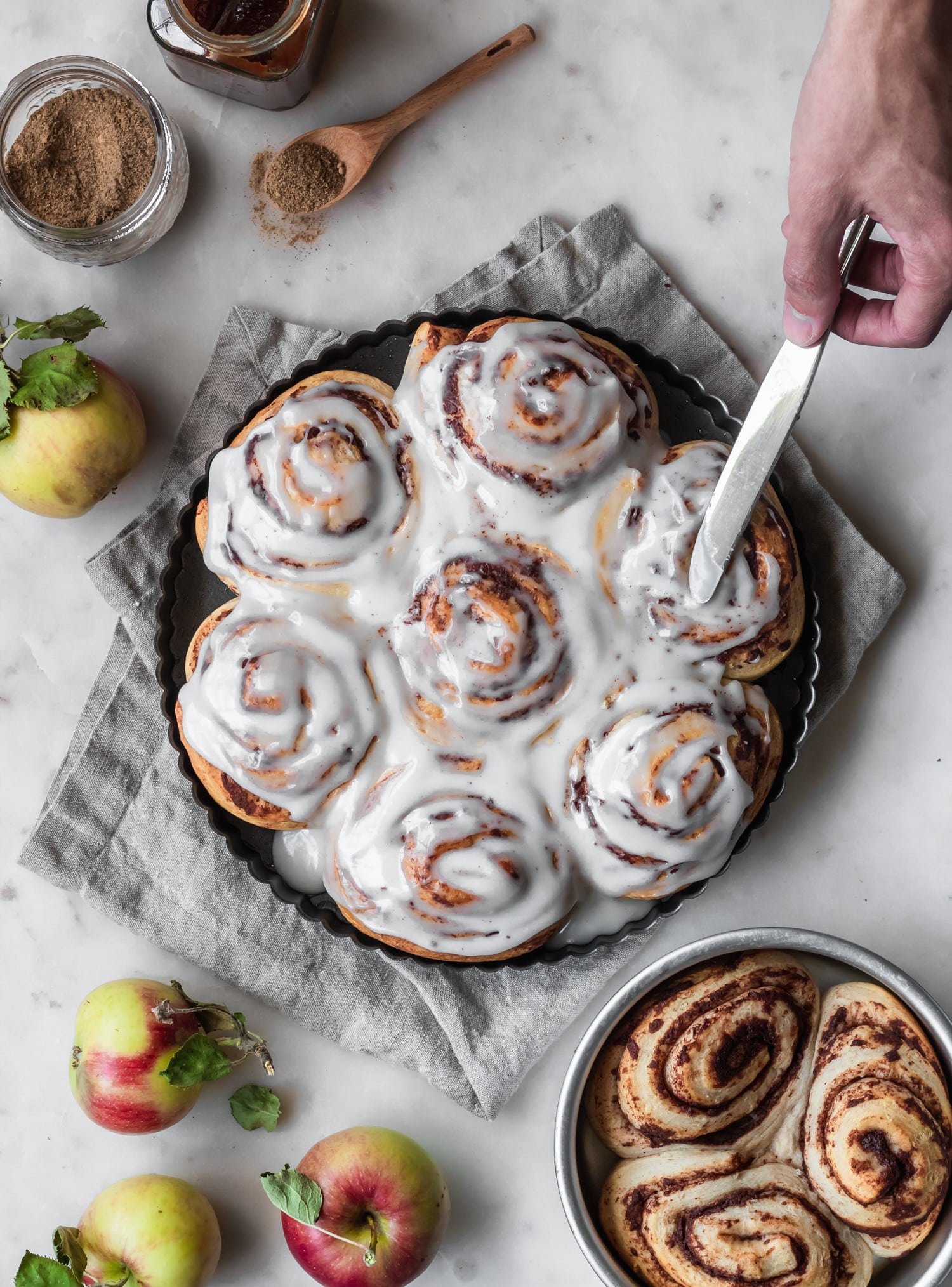 An overhead image of a man spreading brown butter icing over apple butter cinnamon rolls on a marble counter next to a smaller tray of cinnamon rolls, apples, and a grey towel.