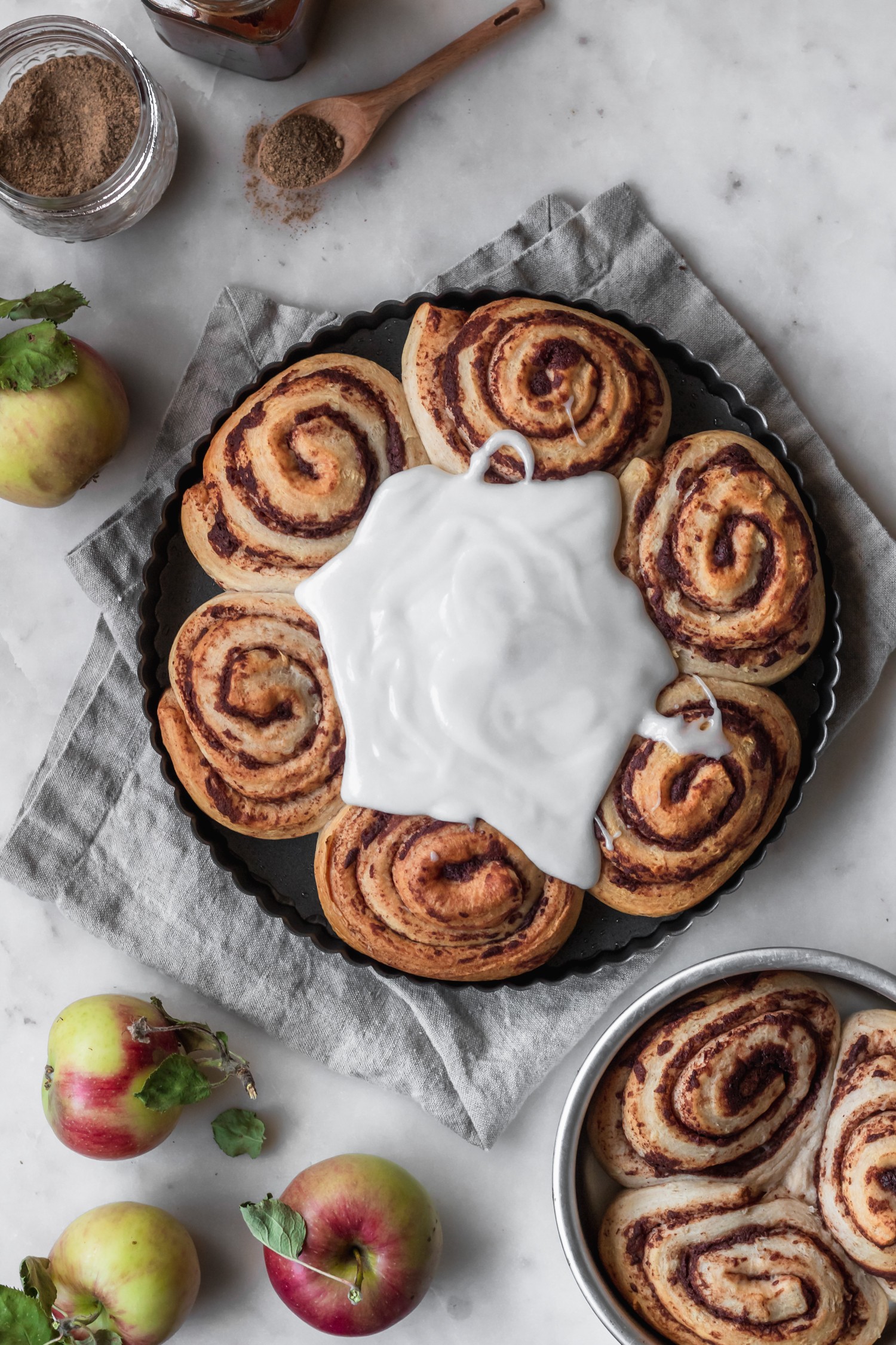 An overhead image of a metal tray of spiced buns with frosting drizzled over them on a marble counter next to a beige linen, small metal tray of buns, and apples.