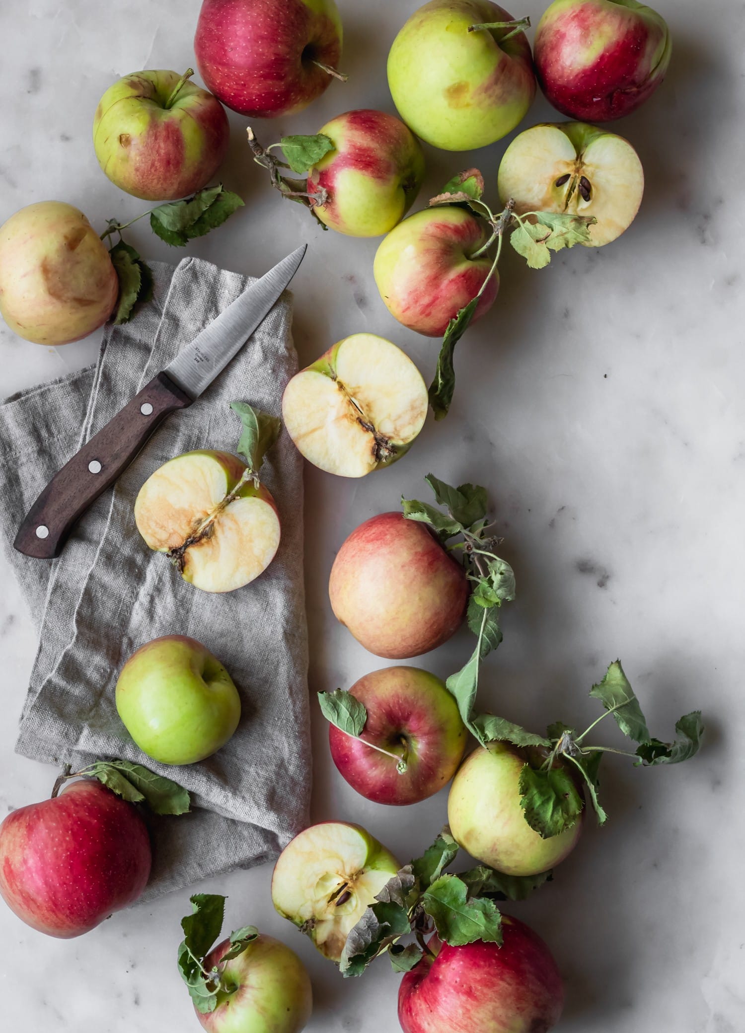 A bird's eye image of apples, some sliced, on a marble counter next to a grey napkin and wood knife.