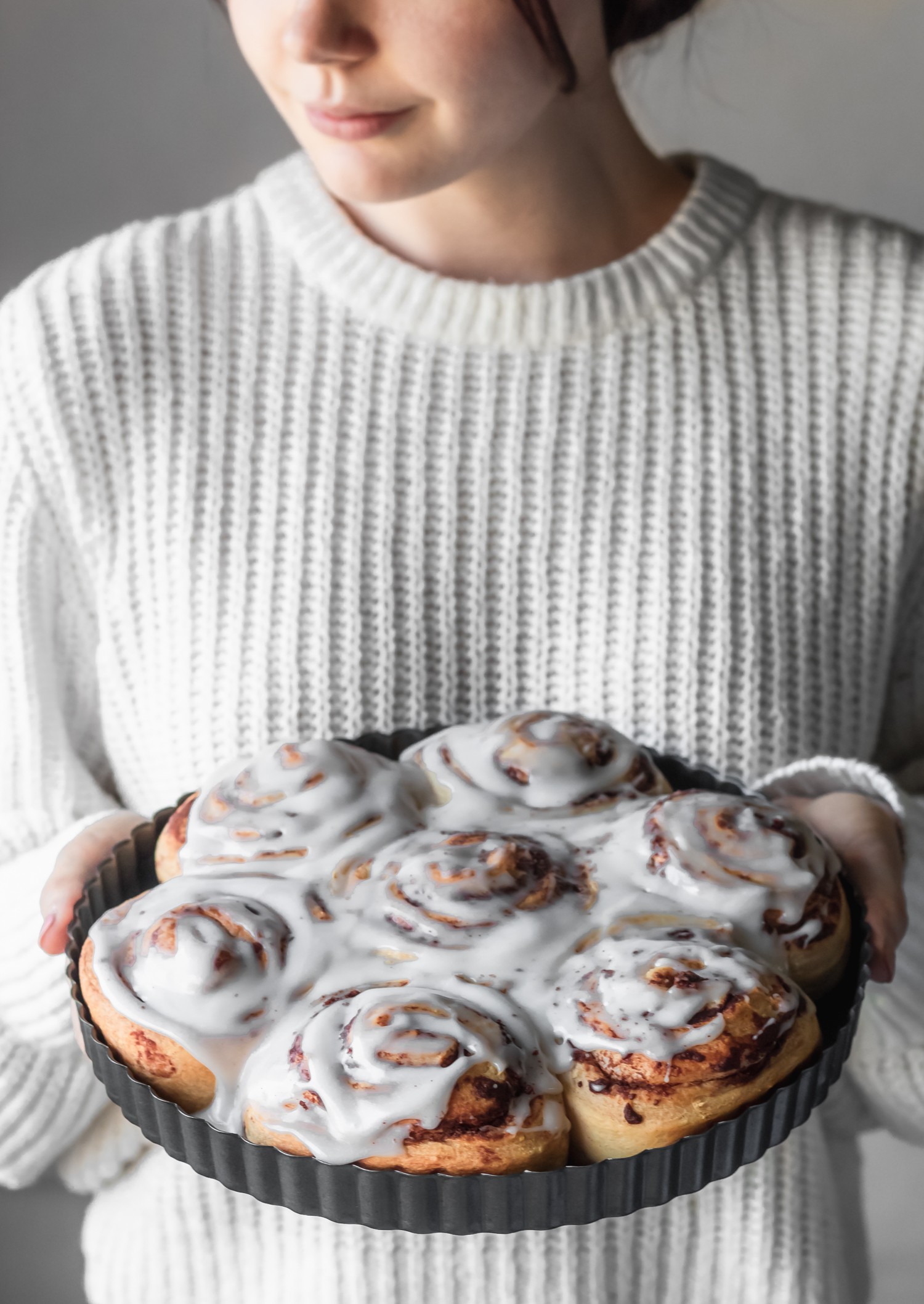 A side image of a woman wearing a white sweater holding a metal tray of iced cinnamon buns.