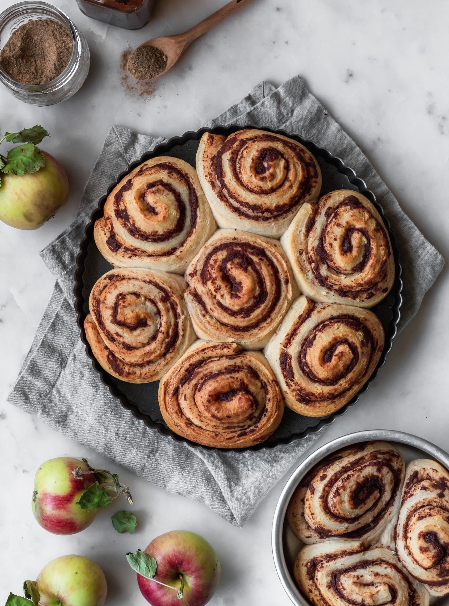 An overhead image of a black dish of morning buns on a white counter next to a grey napkin, smaller tray of buns, and apples.