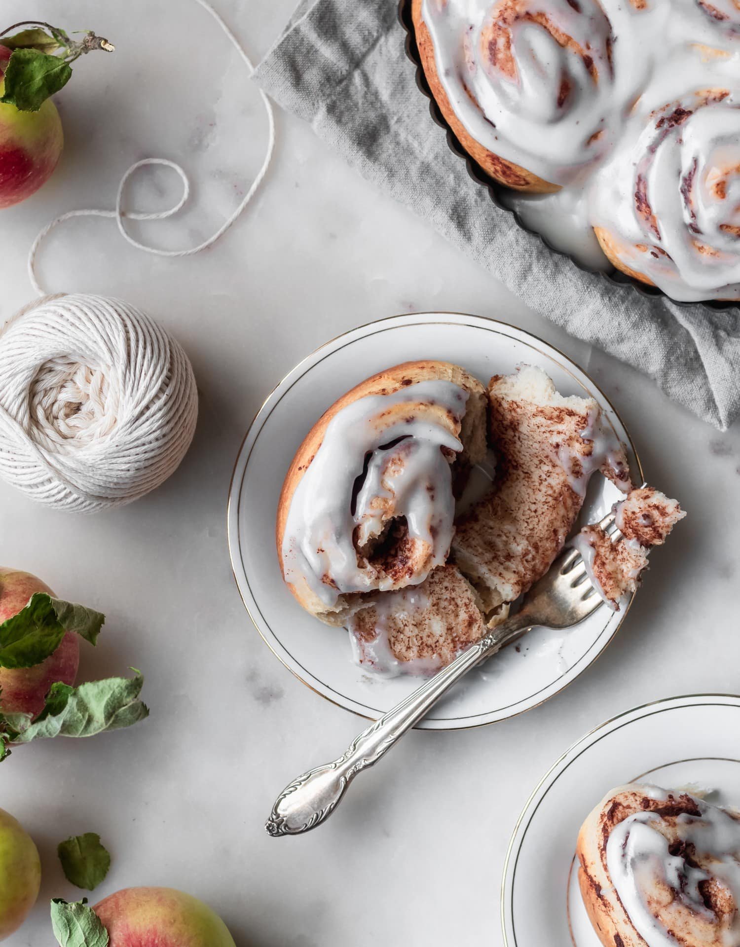 A closeup image of an apple butter cinnamon roll on a white plate with a fork tearing it apart. The plate is on a white table next to another roll and apples.