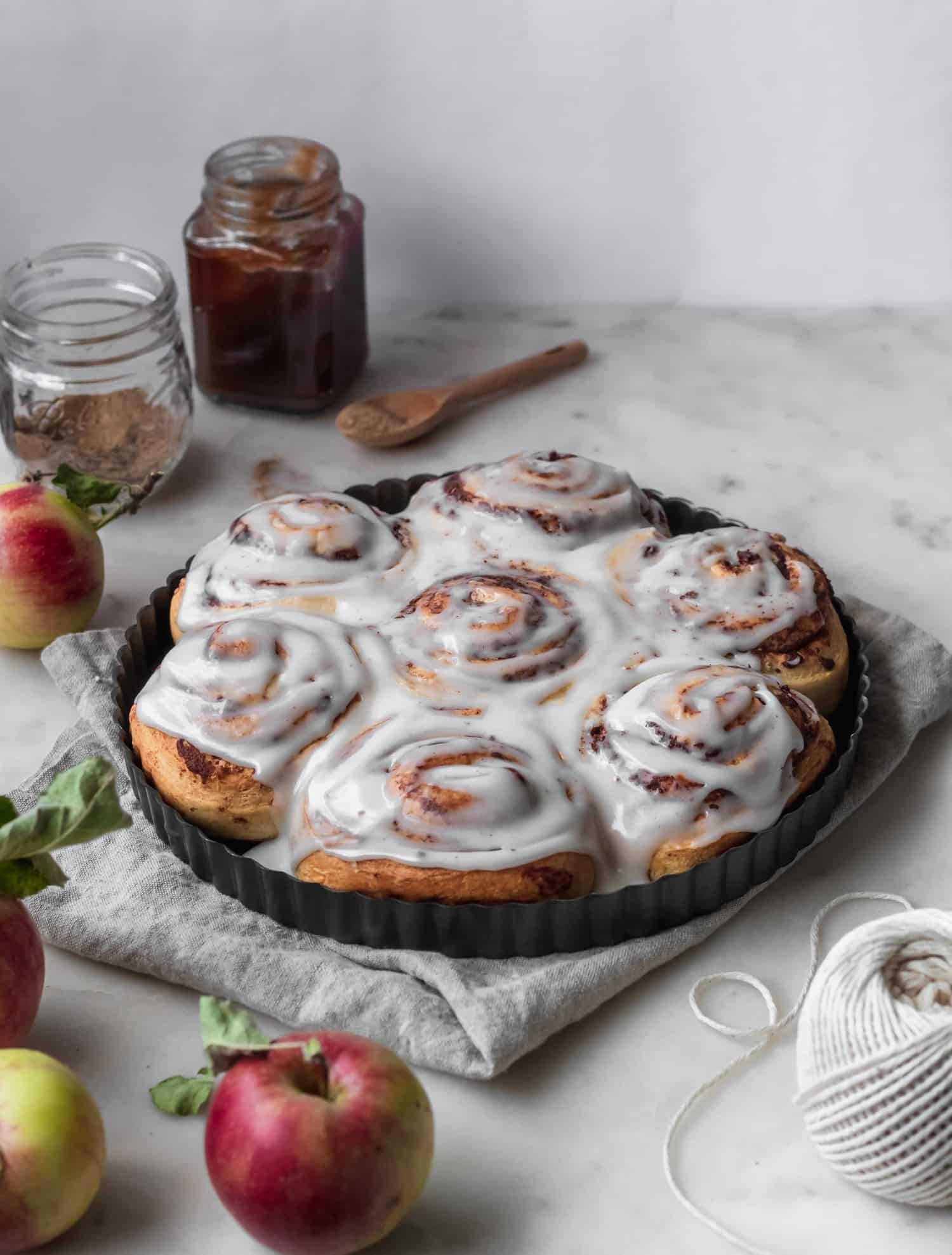 A side image of iced morning buns in a metal baking dish on a white counter next to apples, twine, and a beige linen.