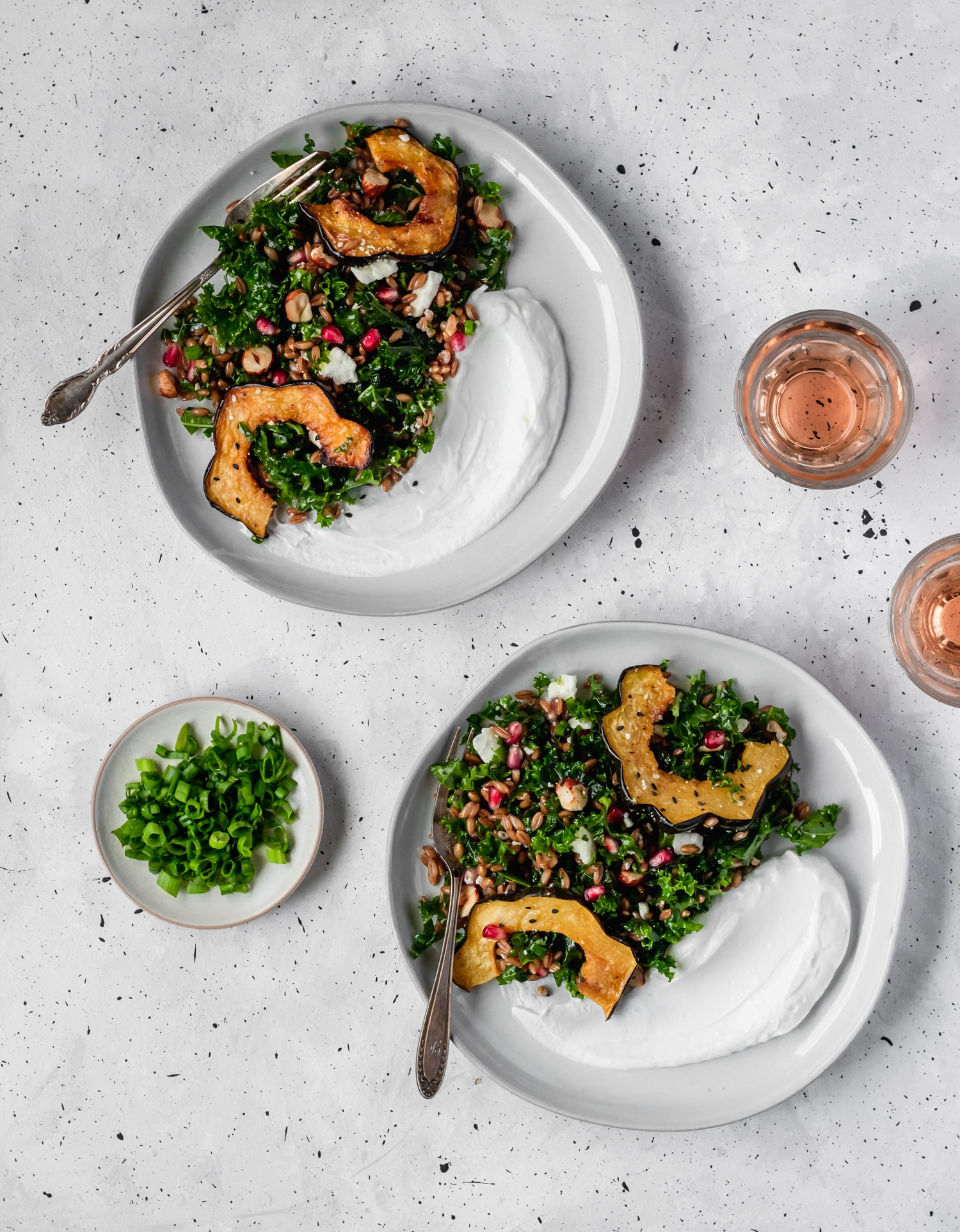 Two white plates of farro salad on a white counter next to a bowl of scallions and two glasses of pink wine.