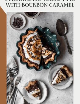 An overhead image of a sliced chocolate cream pie in a dark teal pie plate on a grey speckled table next to two white plates with pie slices, a vintage pie server, vintage forks, a striped linen, and a dish of cacao nibs.
