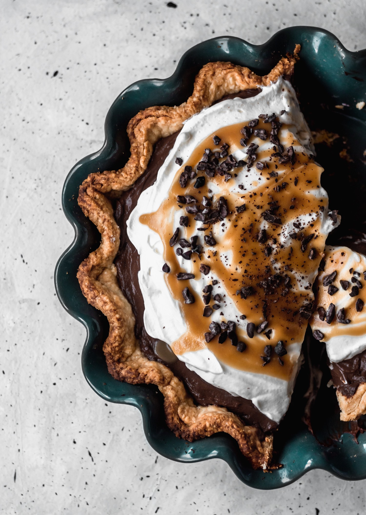 A very closeup image of a chocolate cream pie topped with whipped cream, drizzled with bourbon butterscotch, and sprinkled with cacao nibs.