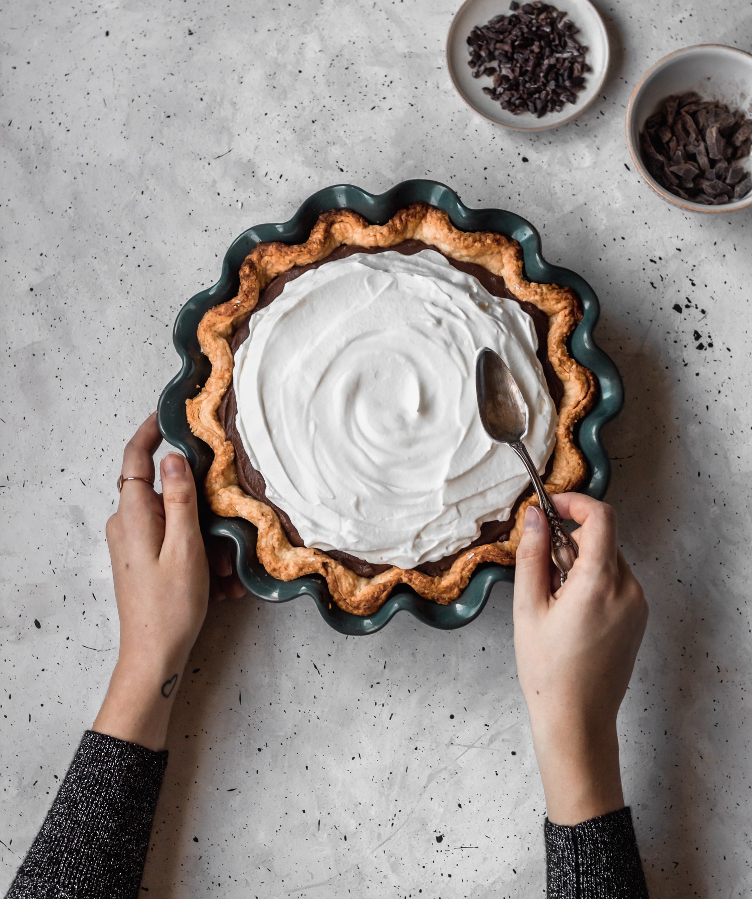 An overhead image of a woman's hands spreading whipped cream over a chocolate cream pie on a grey speckled table.