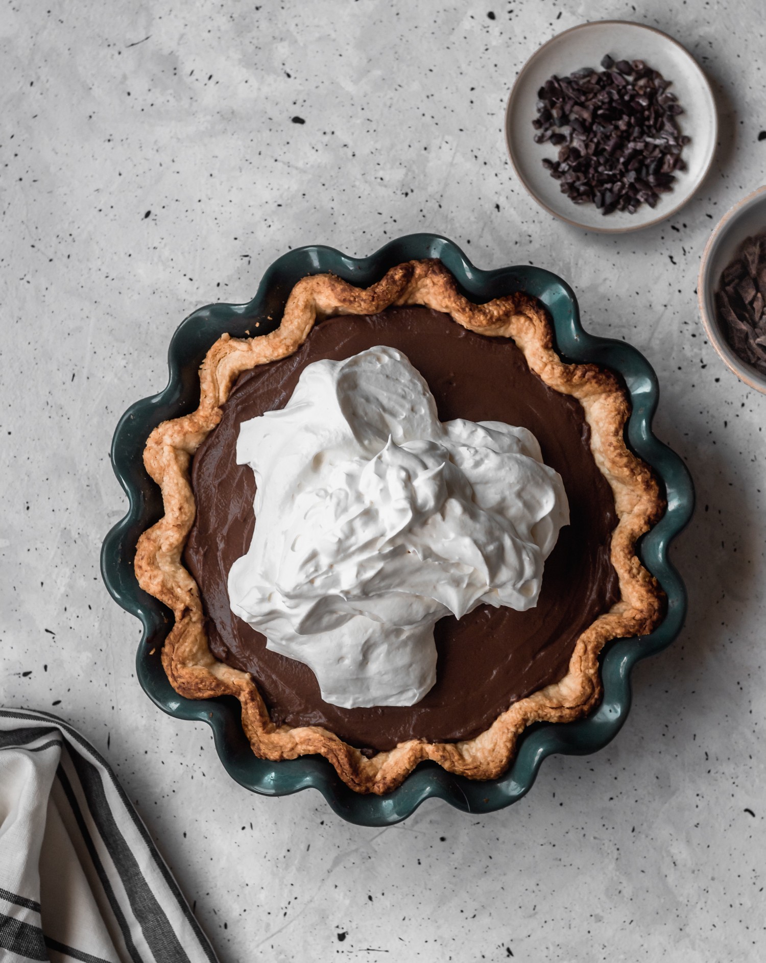 An overhead image of a cocoa tart in a dark teal pie plate topped with a large dollop of whipped cream on a grey table next to a dish of cacao nibs.