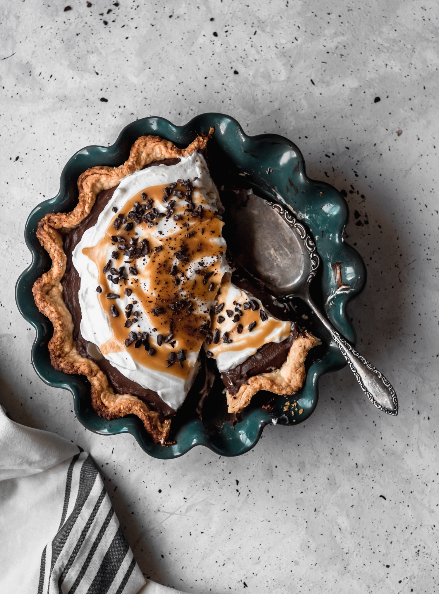 An overhead image of a chocolate cream pie in a dark green pie plate on a speckled grey table. The pie is sliced and there is a vintage pie server in the plate. The dish is next to a striped linen.