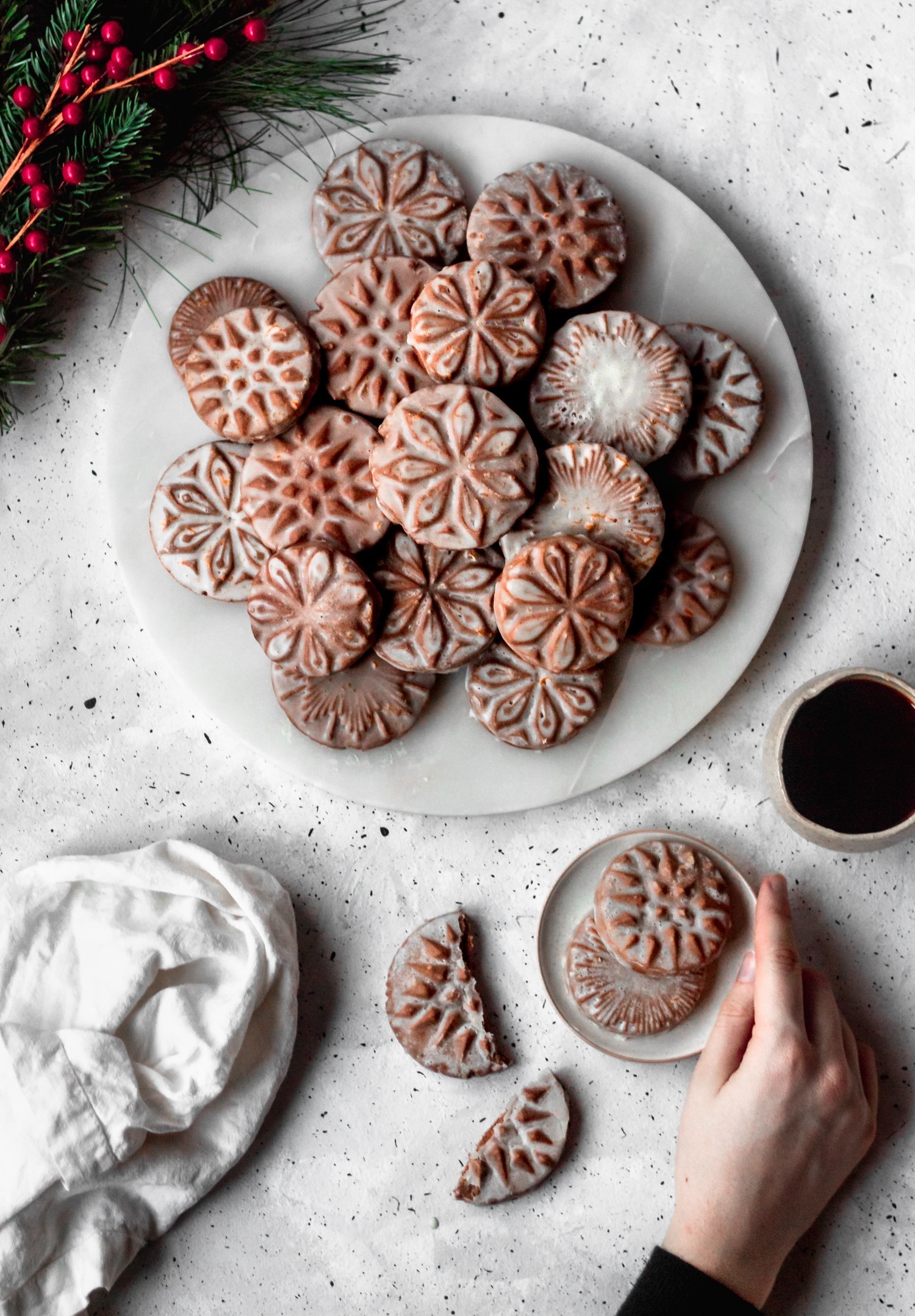 An overhead picture of cardamom gingerbread cookies on a white marble plate on a white table surrounded by pine, a linen, and a cup of coffee.