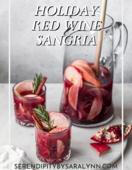 A side image of two glasses of red wine winter sangria garnished with apples, cinnamon, and rosemary on a marble counter next to a cut up pomegranate with a pitcher of sangria in the background.