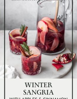 A side image of two glasses of red wine winter sangria garnished with apples, cinnamon, and rosemary on a marble counter next to a cut up pomegranate with a pitcher of sangria in the background.