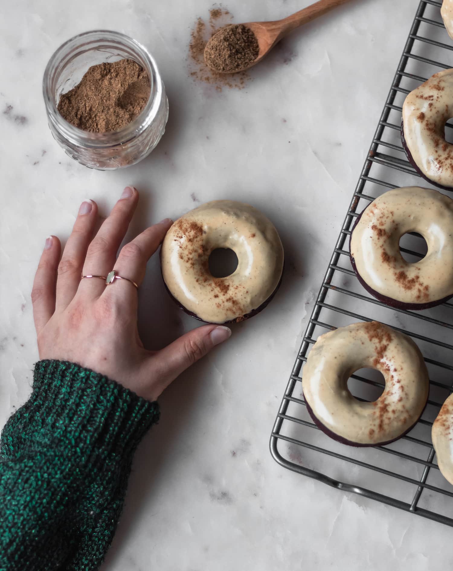 A woman's hand holding a baked chocolate donut on a white counter next to a jar of spices and more donuts.