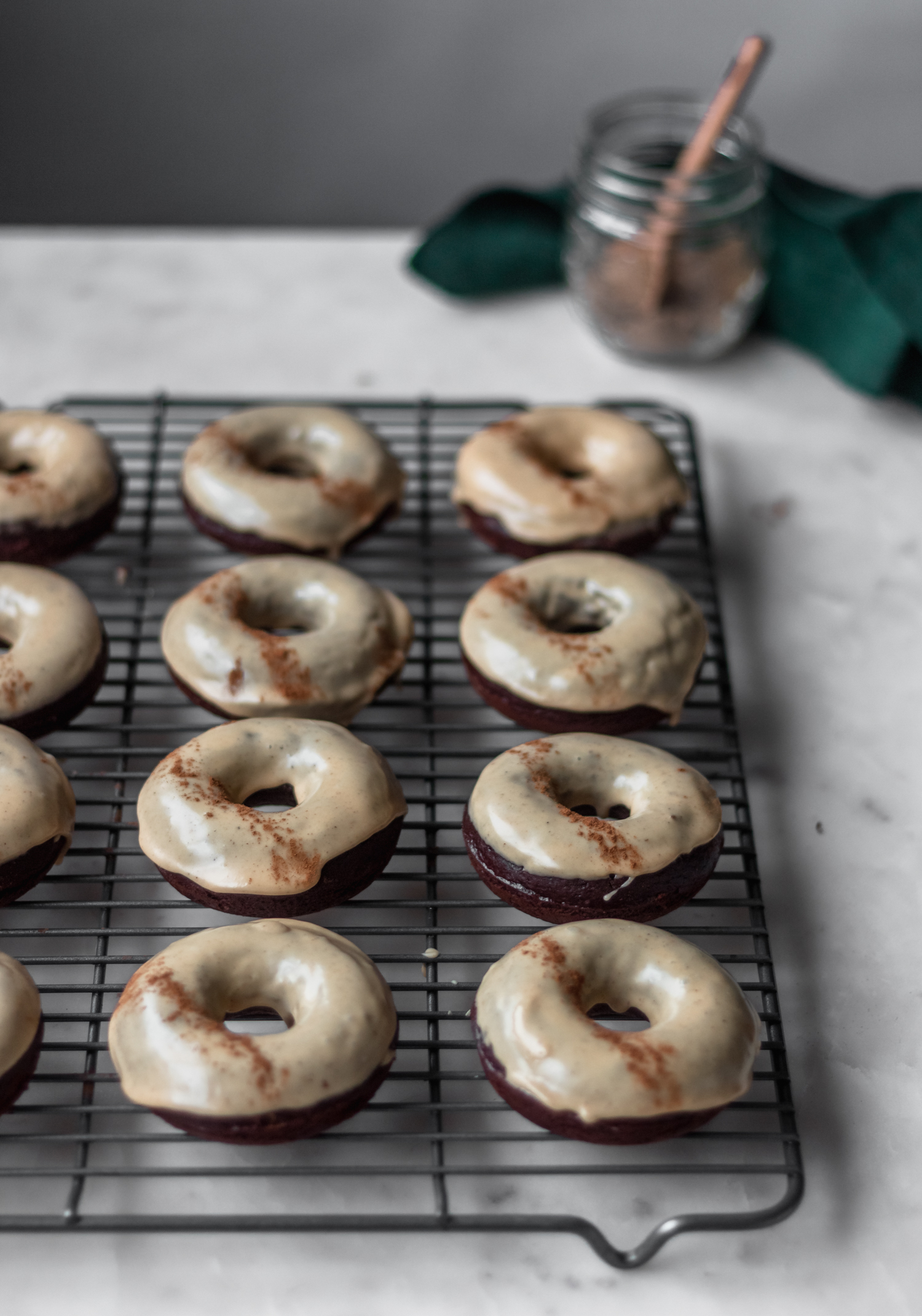 Rows of baked chocolate donuts on a cooling rack on a marble counter with spices and a green linen in the background.