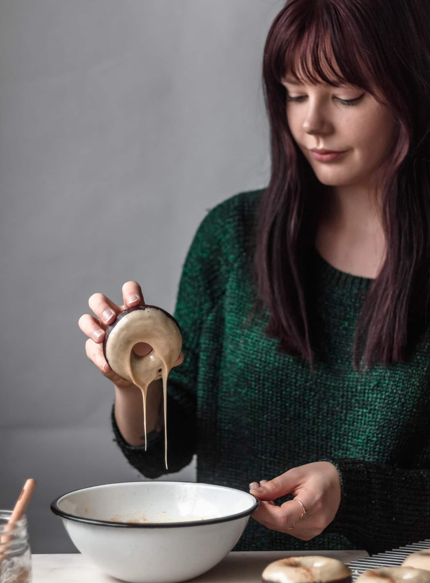 A woman in a green sweater dipping a baked chocolate donut into masala chai glaze on a white counter.