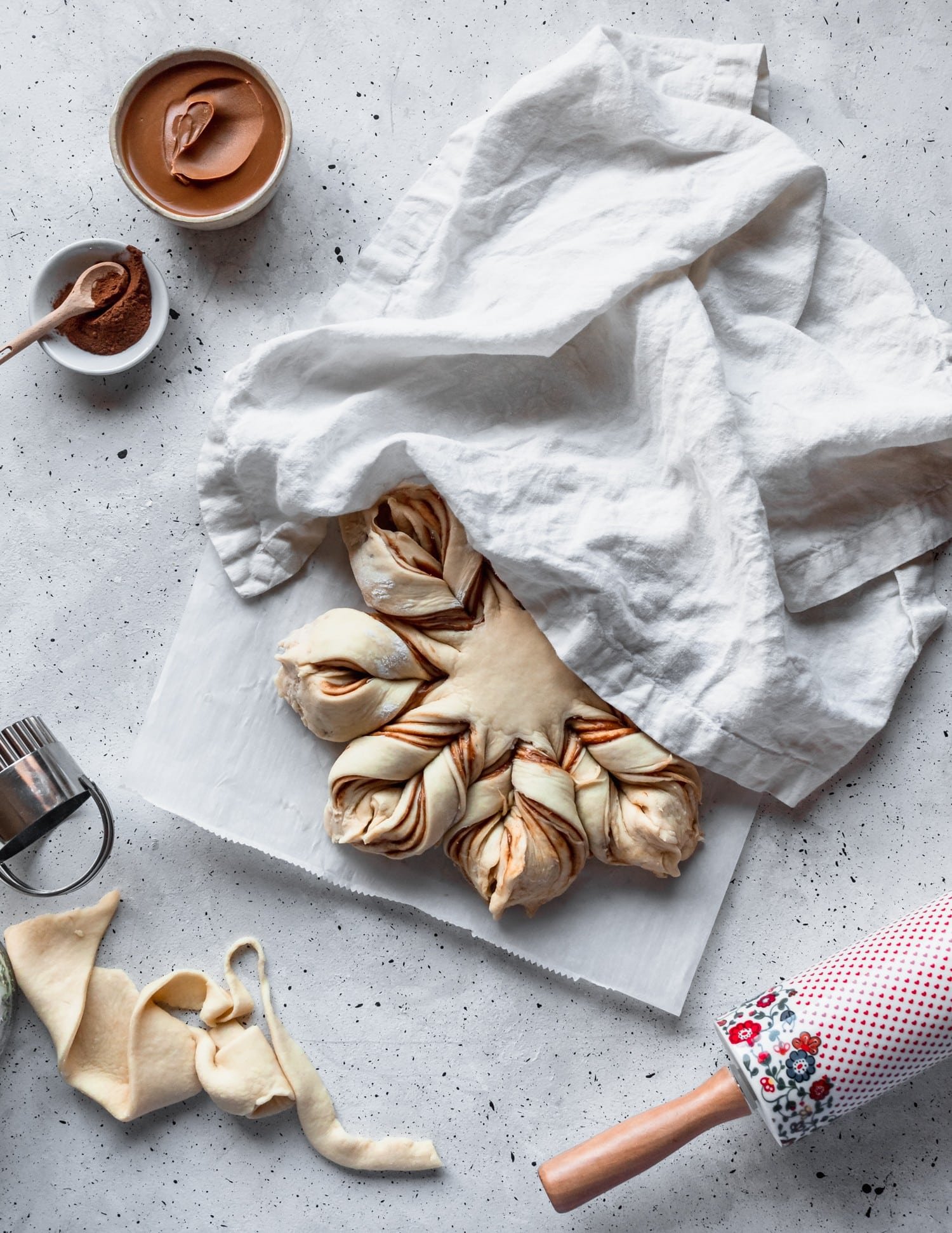 Cookie butter cinnamon star bread rising under a white linen next to a rolling pin on a grey background.