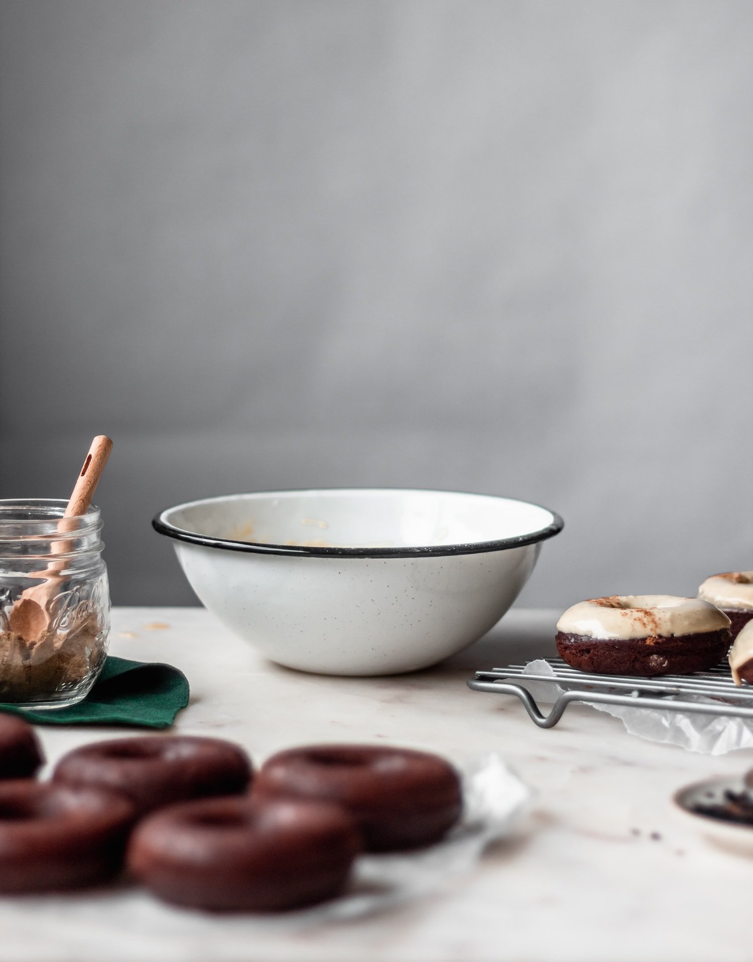 A white bowl of chai glaze on a marble counter next to a jar of spices and donuts.