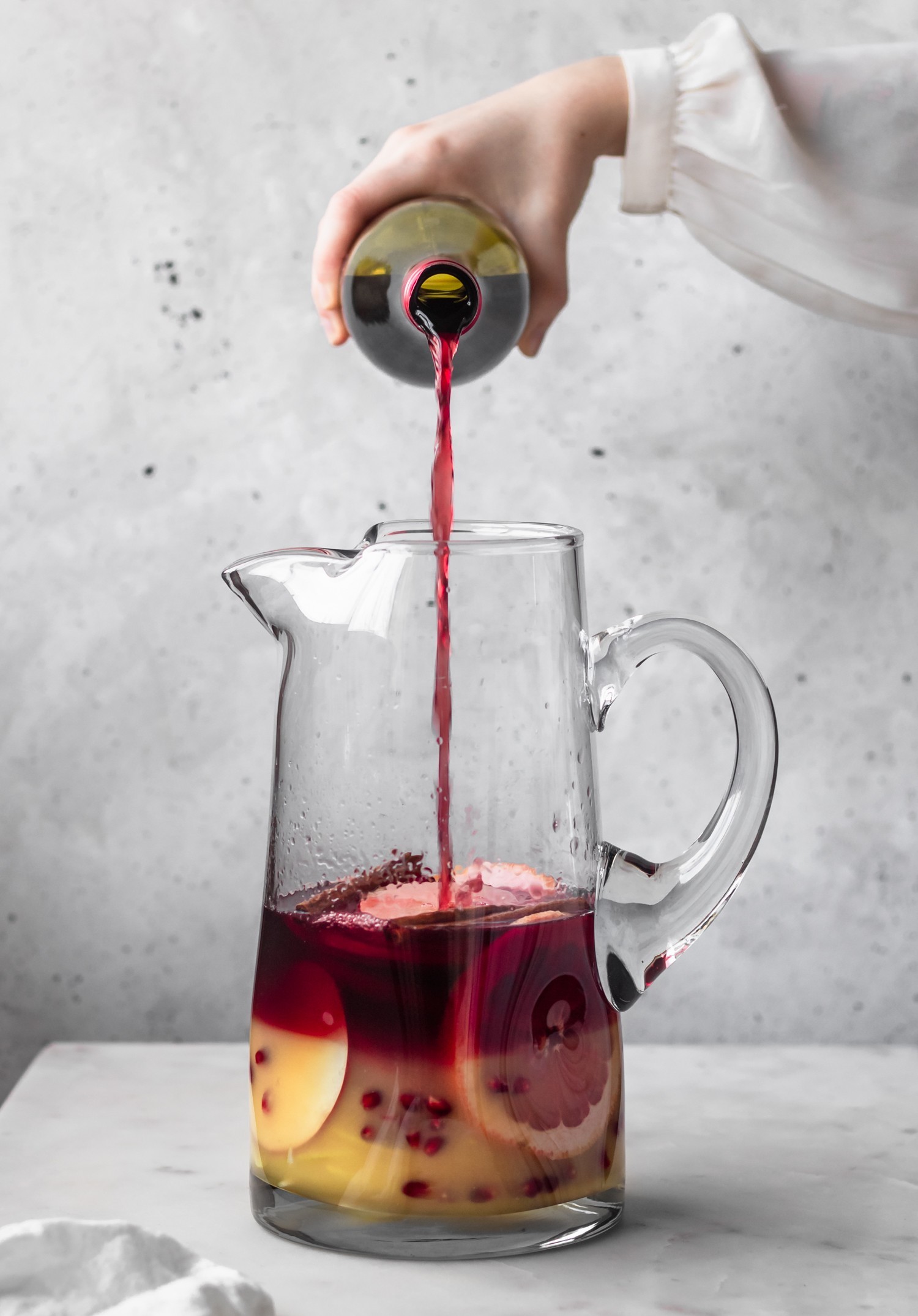 A side image of a woman's hand pouring red wine into a pitcher on a marble counter with a grey background.