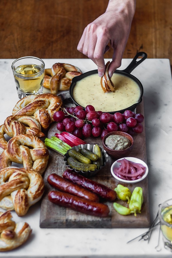 A man's hand dipping a soft pretzel into beer cheese fondue, surrounded by fruits, pickled veggies, beer, and pretzels.