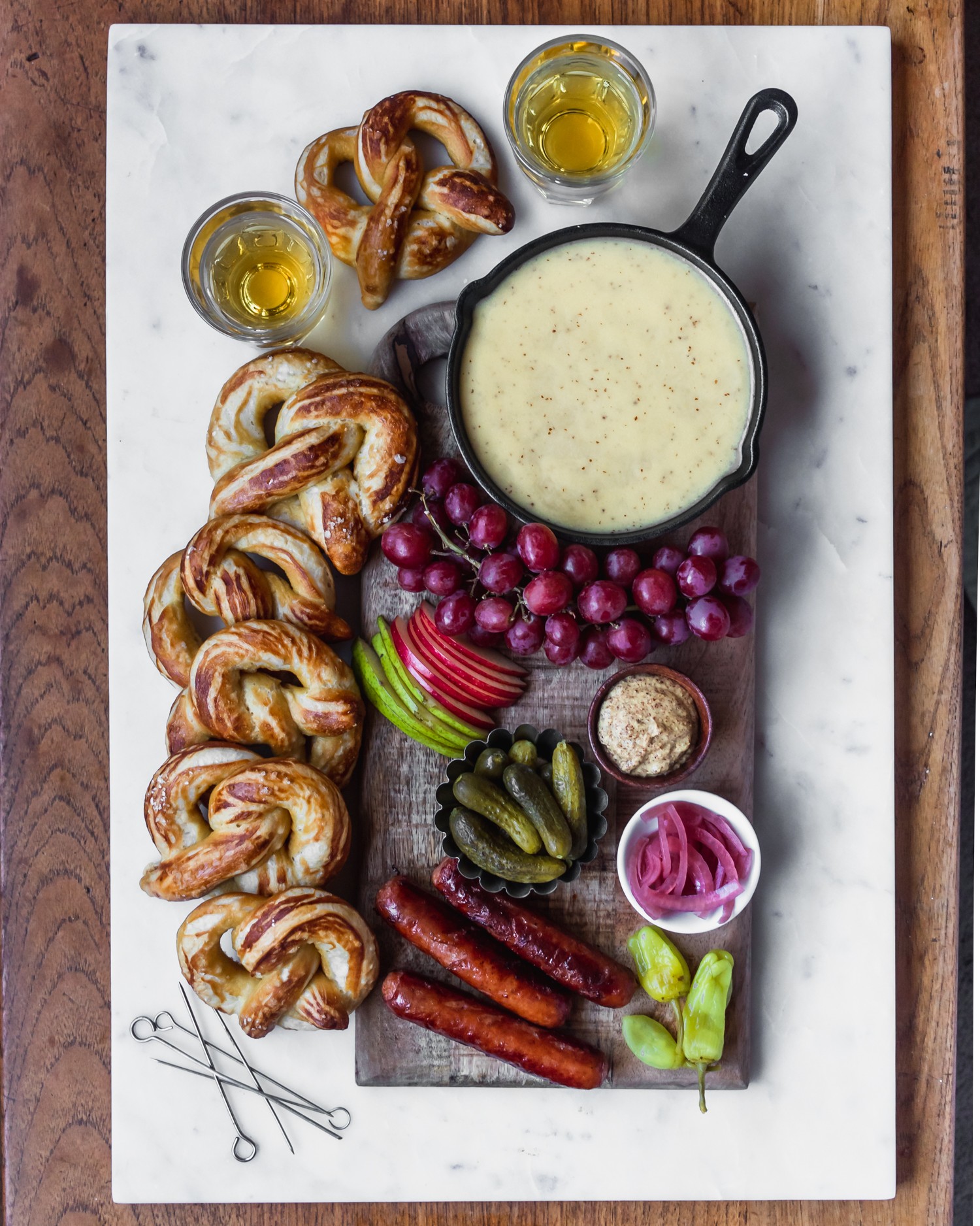 A skillet of beer cheese fondue surrounded by soft pretzels, fruit, pickled vegetables, brats, and beer.