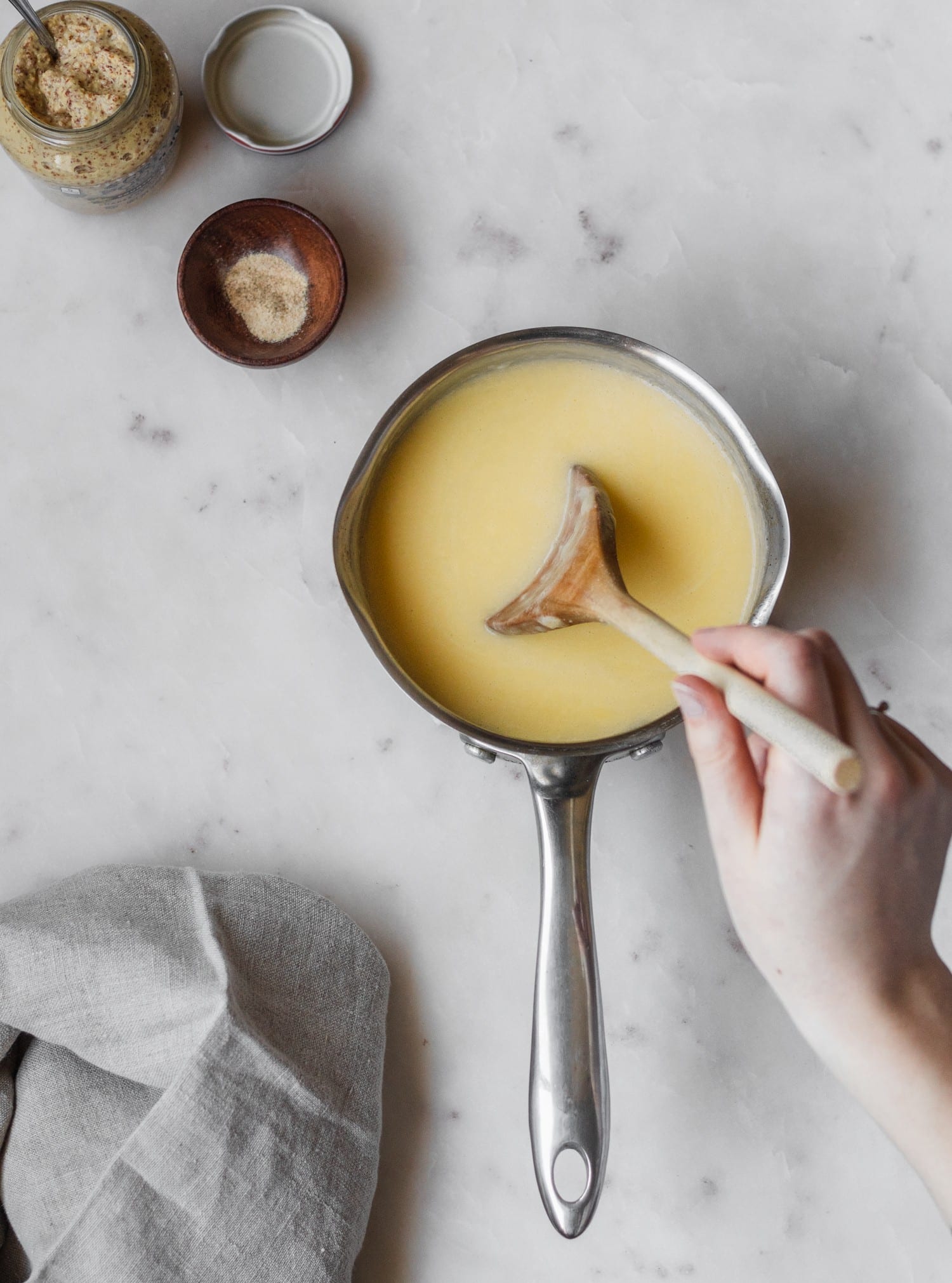 A woman's hand stirring a pot of cheese dip sitting on a white marble background. The pot is sitting next to a jar of Dijon and spices.