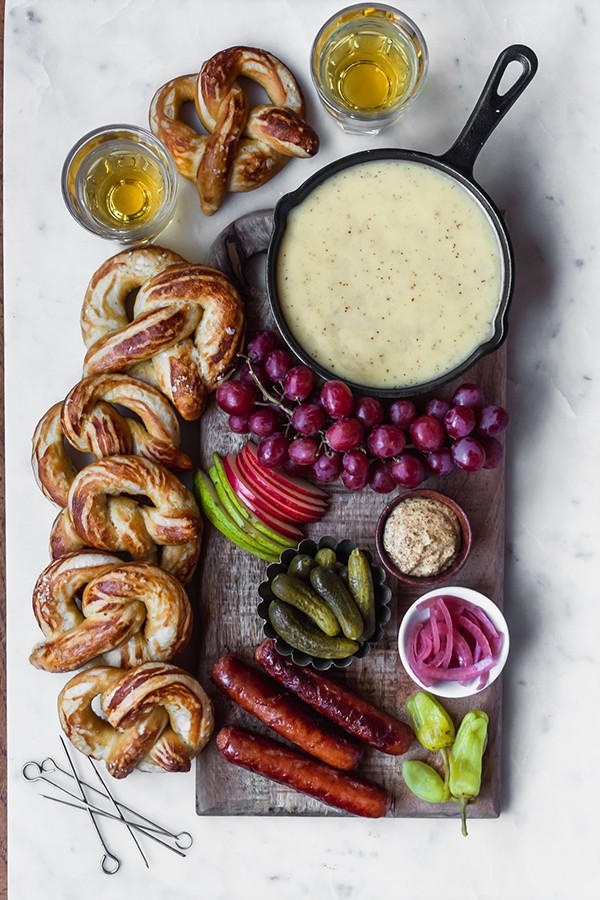 Beer cheese fondue in a skillet on a wooden board sitting on a white table, surrounded by fruit, vegetables, pretzels, and beer.