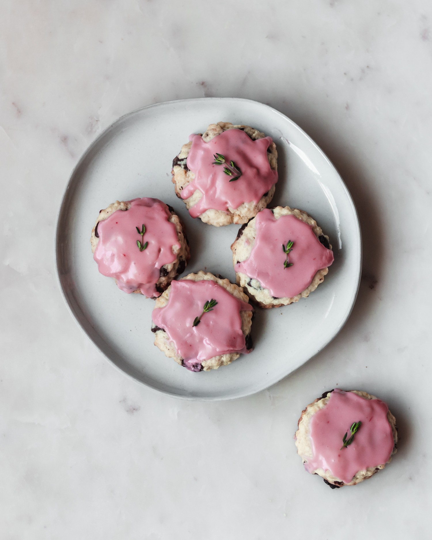Four chocolate chip scones with pink glaze on a white plate sitting on a marble background.