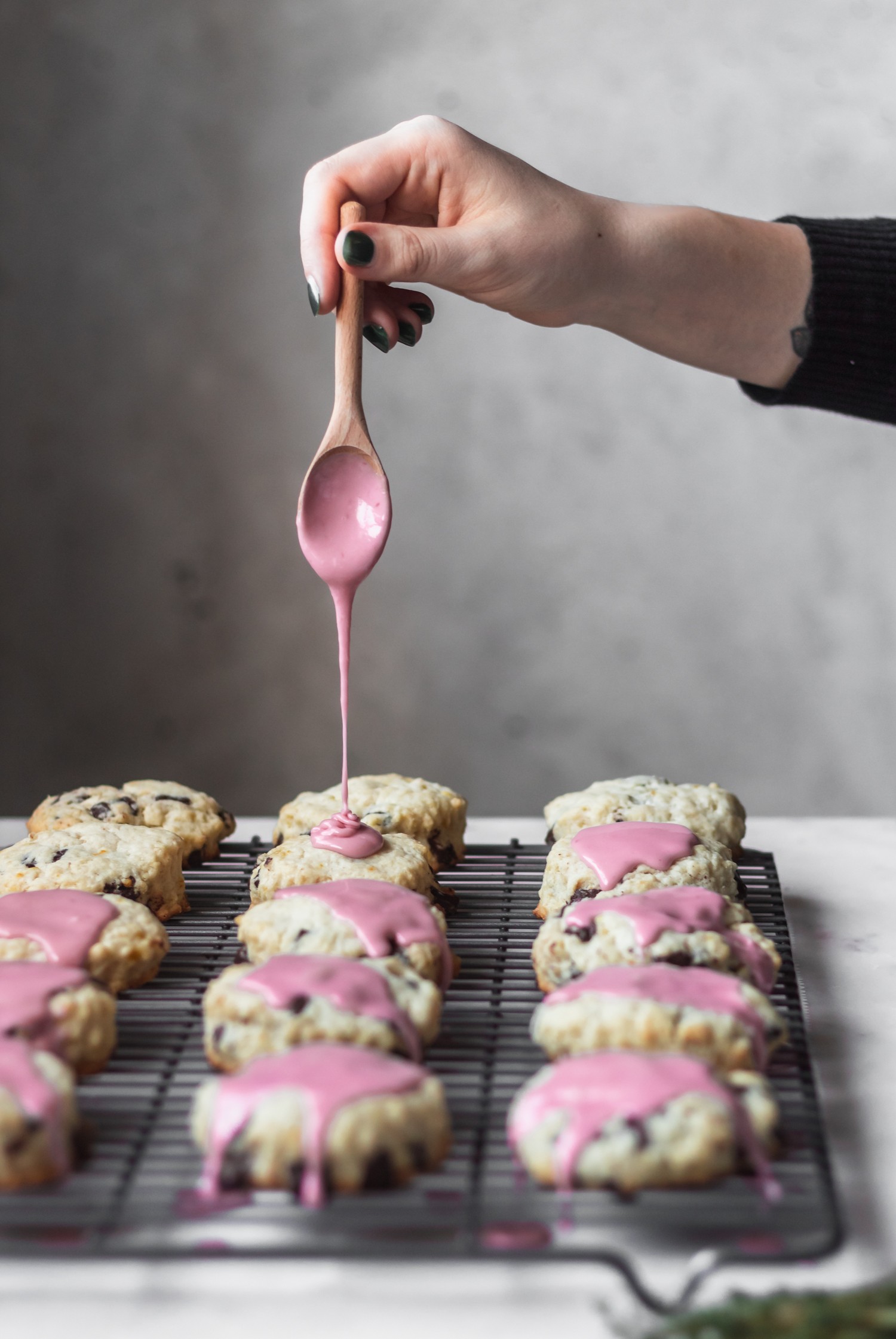 Chocolate biscuits lined on a wire rack while a woman's hand drizzles pink glaze on top.