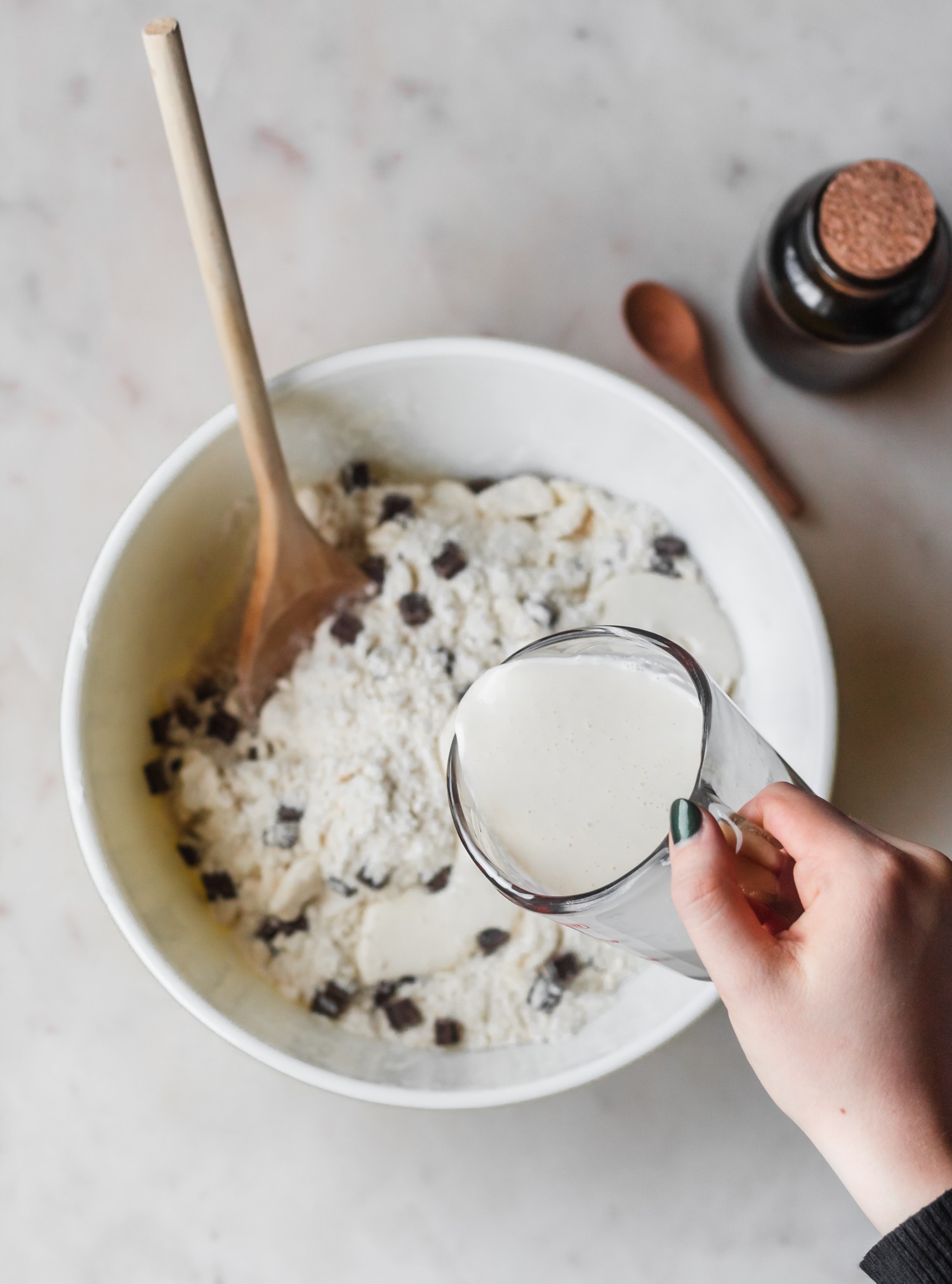 A white bowl filled with flour and chocolate chunks. There is a wooden spoon in the corner, and a woman's hand is pouring cream into the bowl. The bowl is on a white surface, and there is a jar of vanilla extract in the corner.