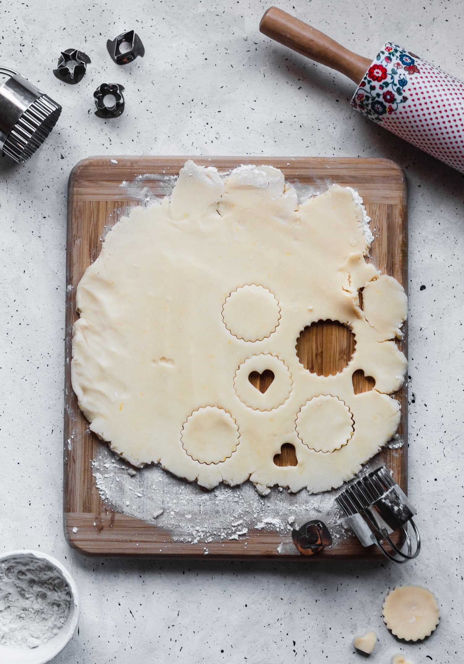 Rolled-out dough on a wooden board, surrounded by cookie cutters, flour, and a rolling pin.