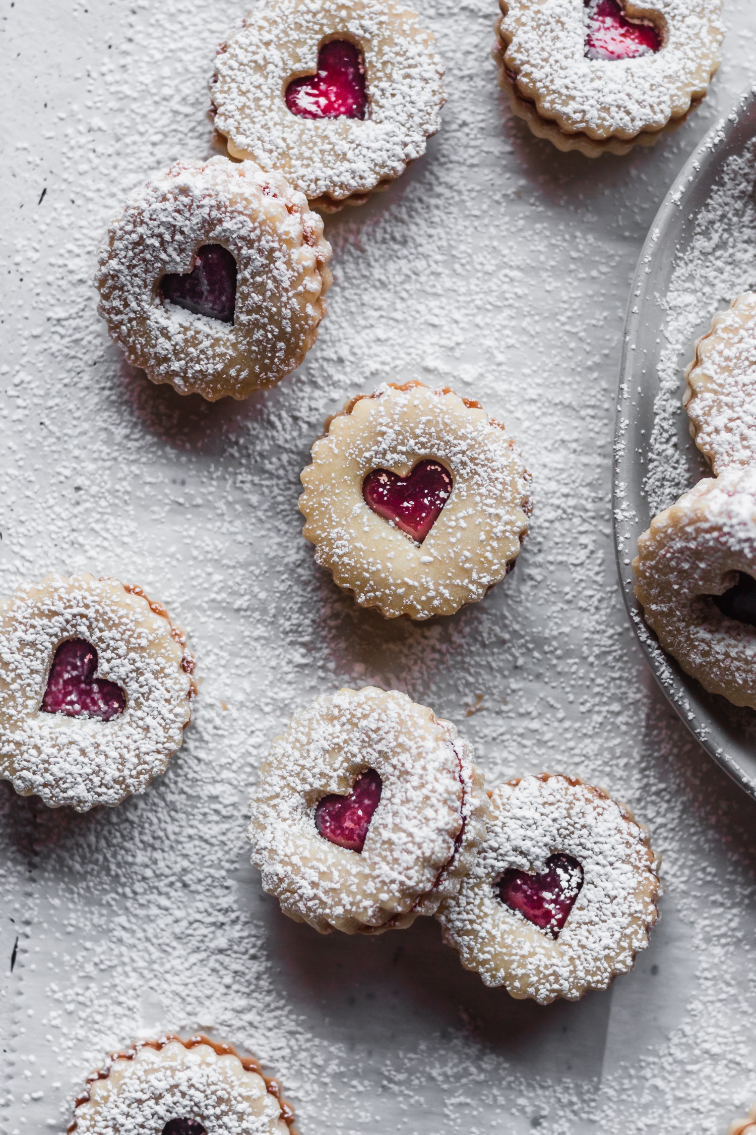 Piles of shortbread linzer cookies with raspberry jam on a white counter next to a white plate of cookies.