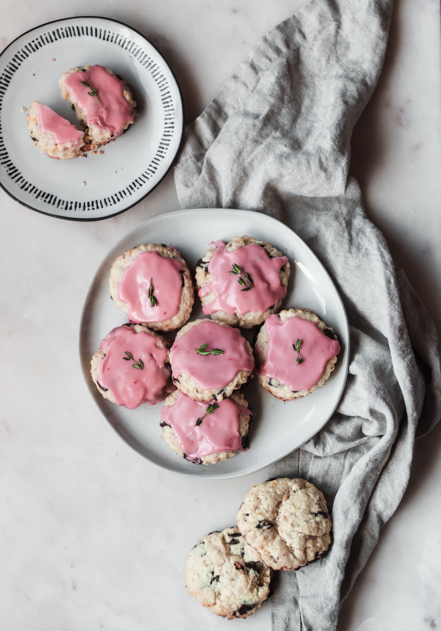 A white plate of chocolate chip scones drizzled with blood orange glaze. There is a beige linen across the marble table with two unglazed scones in the corner and a small plate with a scone in the upper left corner.