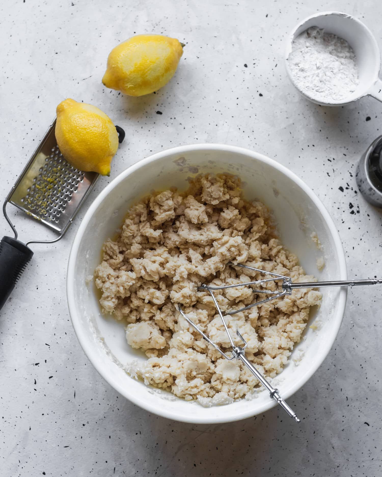 A white bowl filled with crumbly dough, surrounded by lemons, flour, and a jar of vanilla extract.