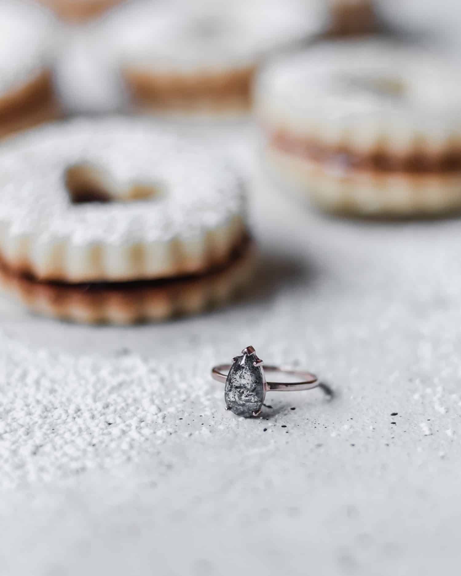 Salt and pepper diamond, pear-shaped ring with rose gold band on a grey surface with linzer cookies in the background.