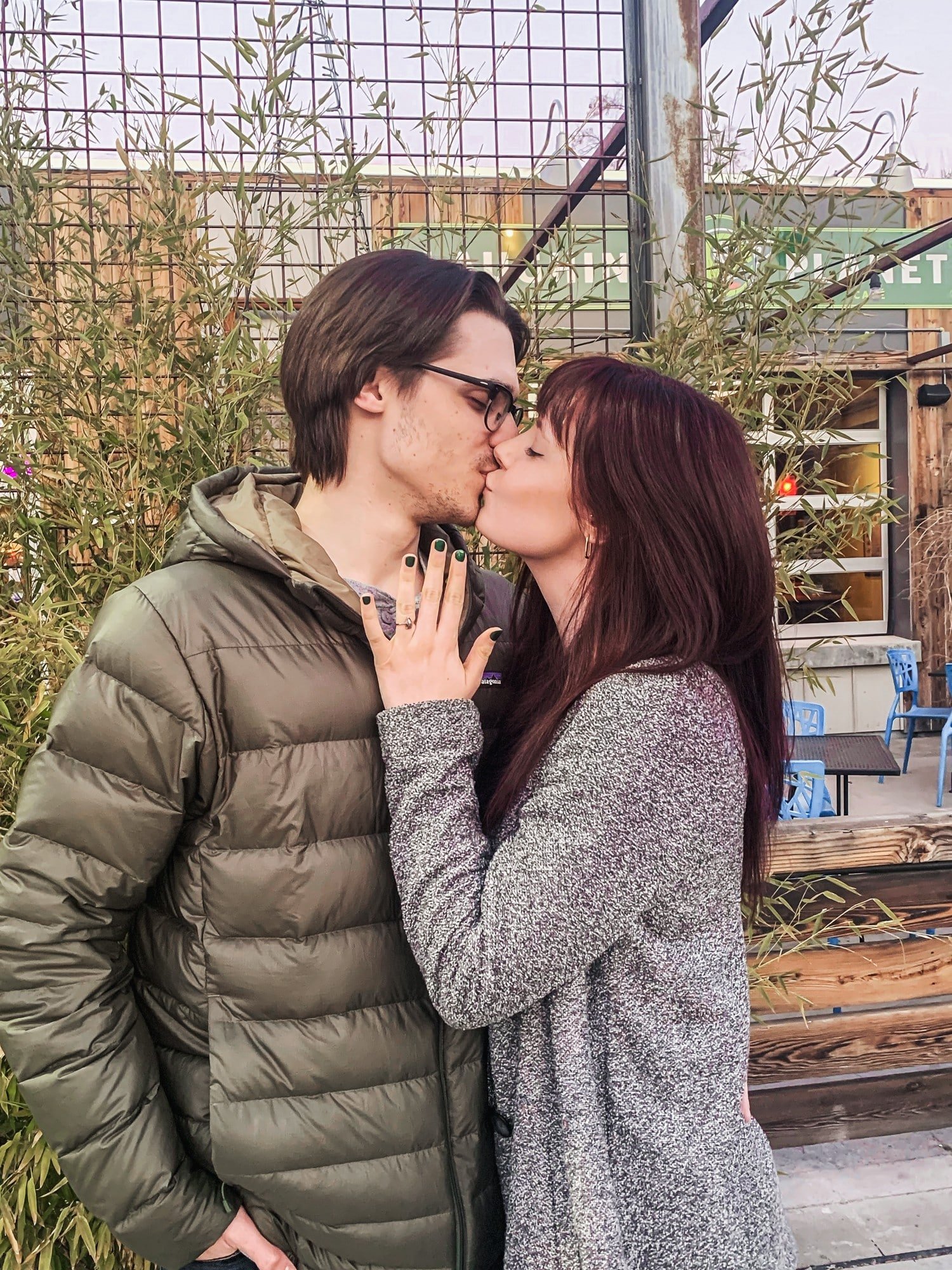 Man and woman standing in front of a green tree kissing. Woman is holding up hand with engagement ring.