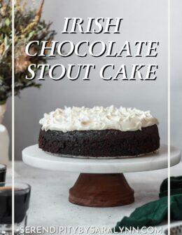 A side image of a chocolate stout cake with Irish cream cheese frosting on a wood and marble cake stand on a white counter next to a green linen, cup of stout, and bouquet of flowers.
