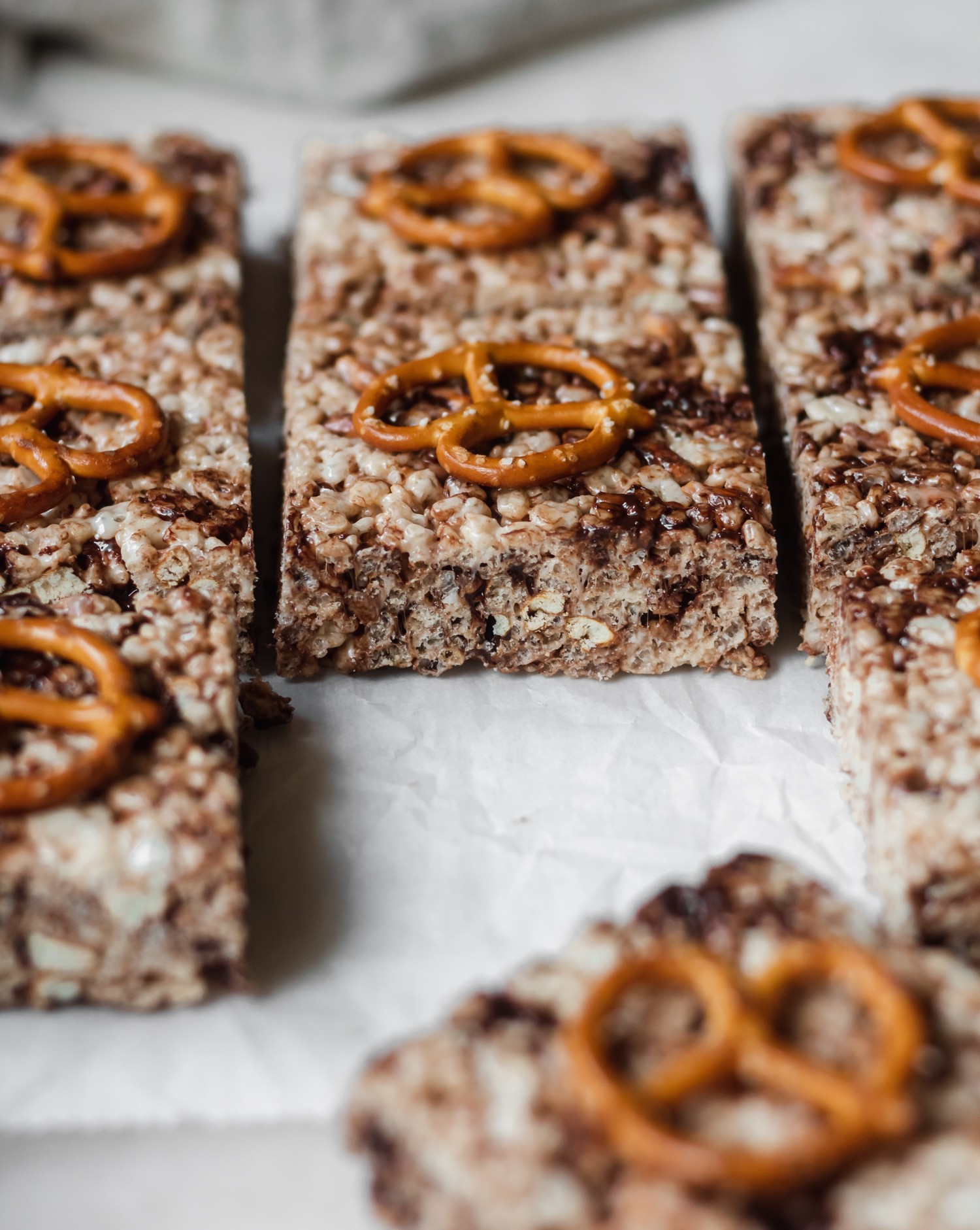 A closeup of cereal bars with chocolate and a pretzel on top. Cereal bars are on white parchment paper.