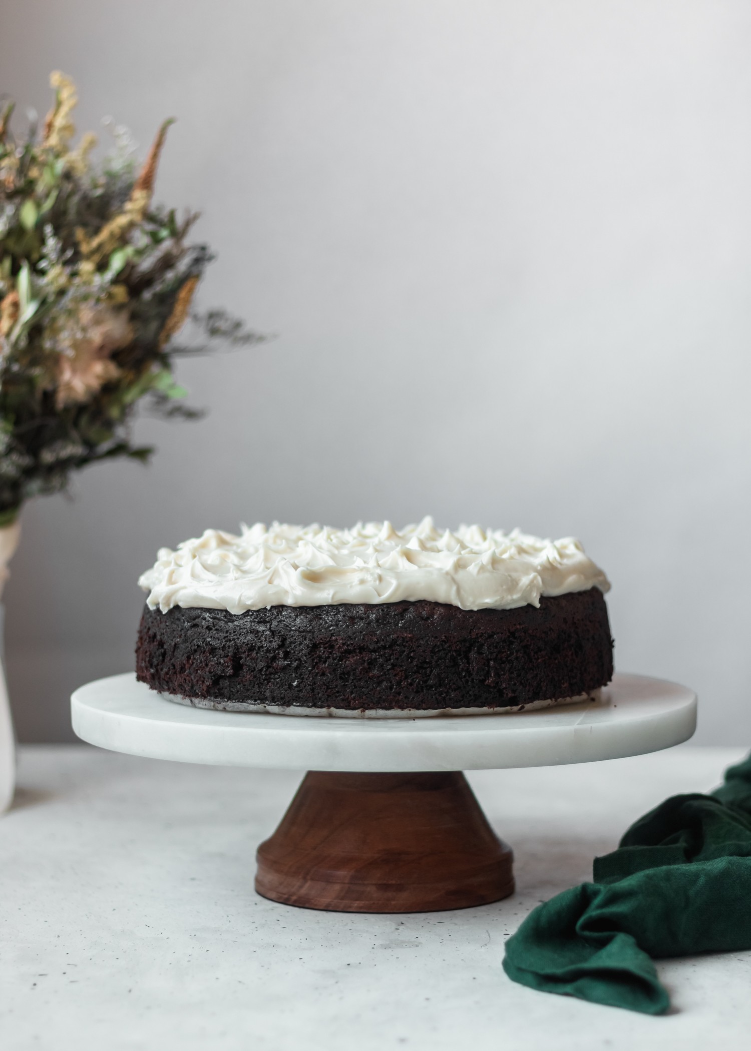 Stout cake on a marble and wood stand against a white background with a bouquet of flowers on the left side and a green linen on the right side.