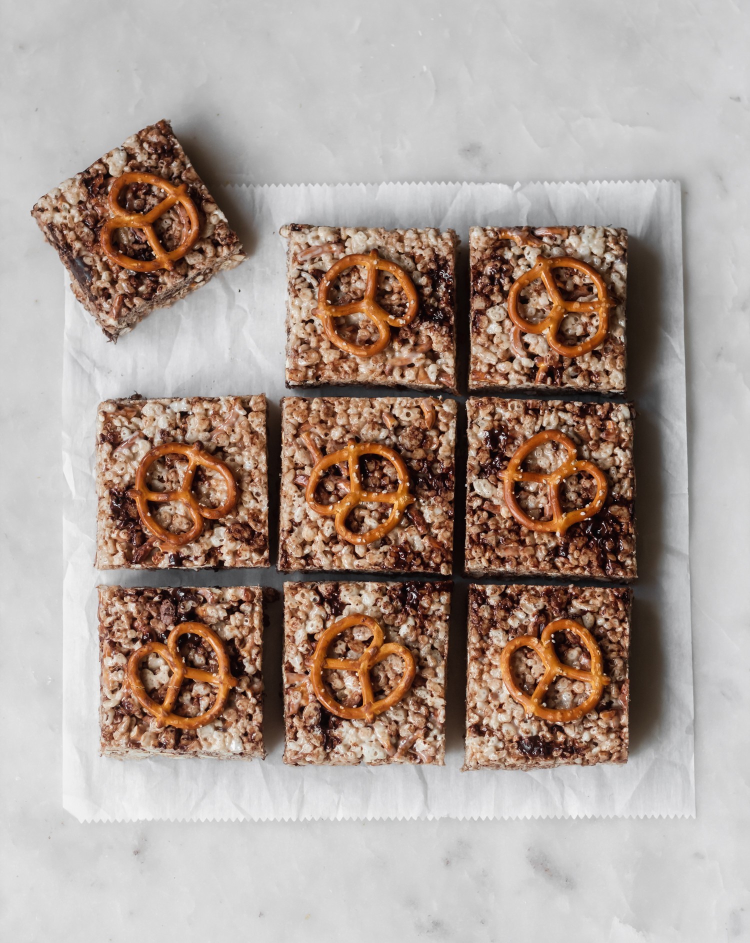Cut chocolate rice krispie treats with pretzels. One in the left corner is pulled away. Cereal bars are sitting on white parchment paper.