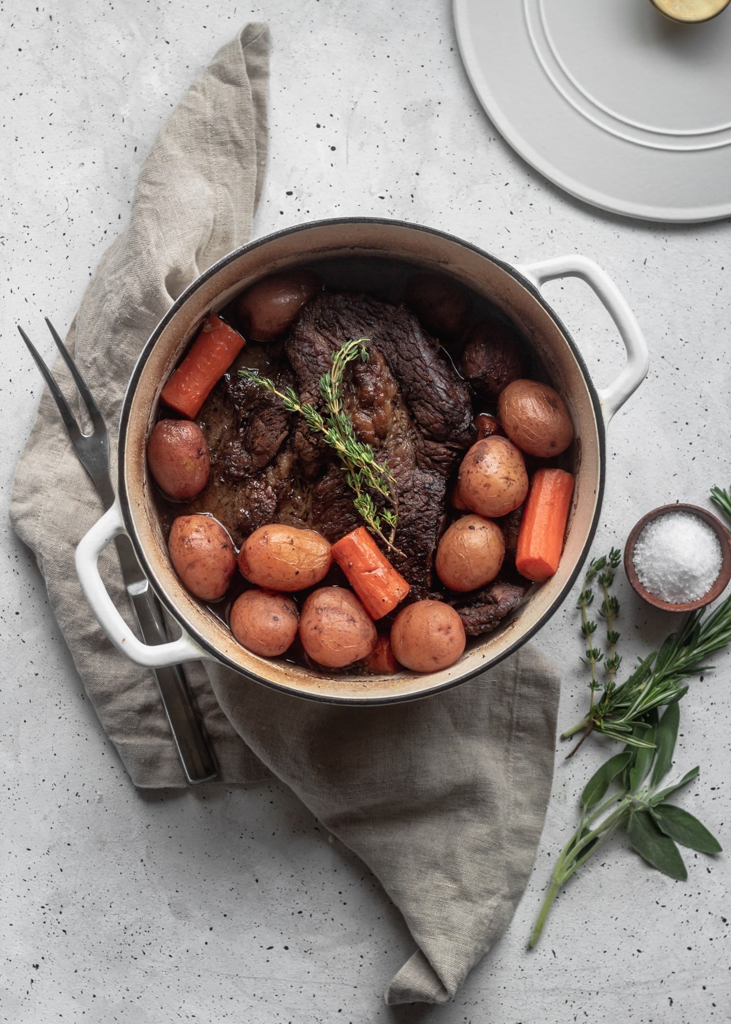 A white pot filled with beef, potatoes, and carrots with a sprig of thyme on top. The pot is sitting on a beige towel on a white background with herbs in the right bottom corner.