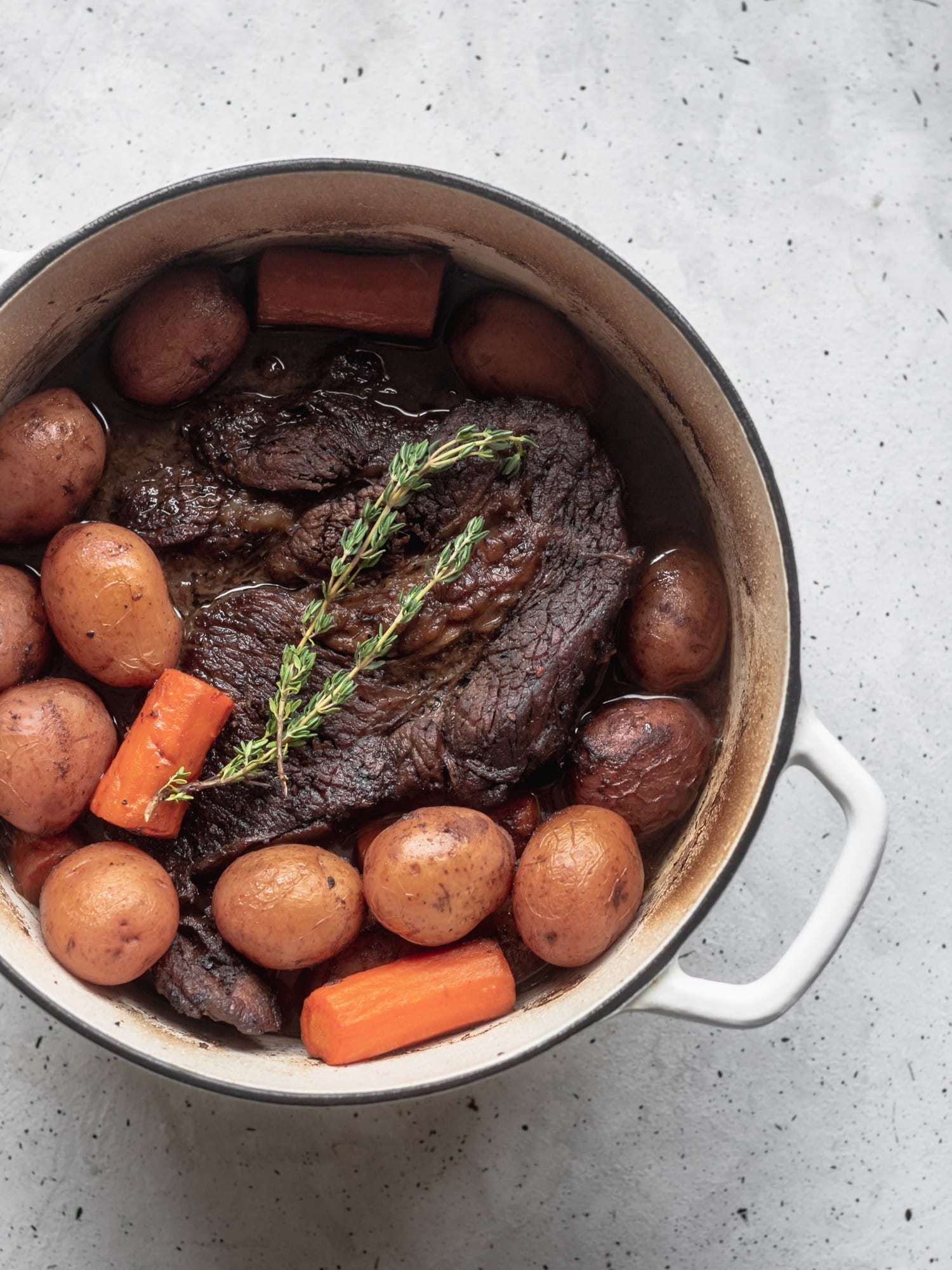 A closeup shot of a Dutch oven filled with pot roast, topped with a sprig of thyme.