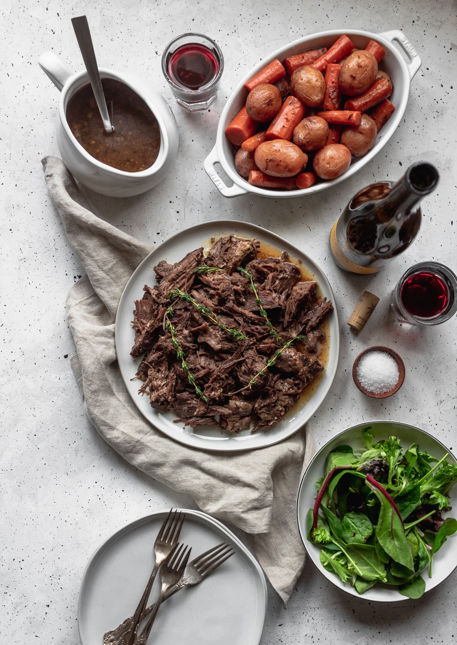 A platter of pot roast on a white background surrounded by a plate of vegetables, a bowl of salad, gravy, and red wine.