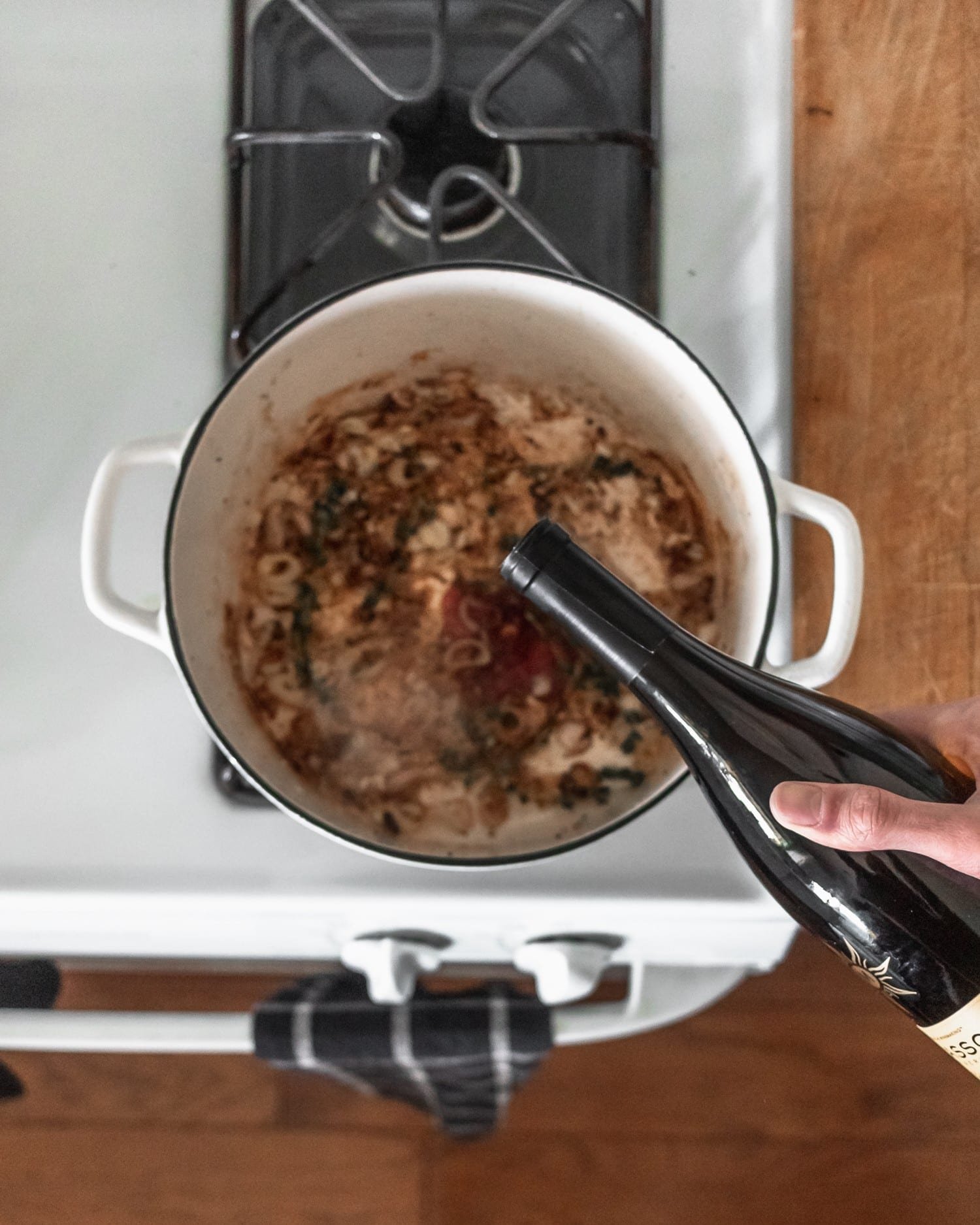 A man's hand pouring red wine into a pot of onions on a white stove.