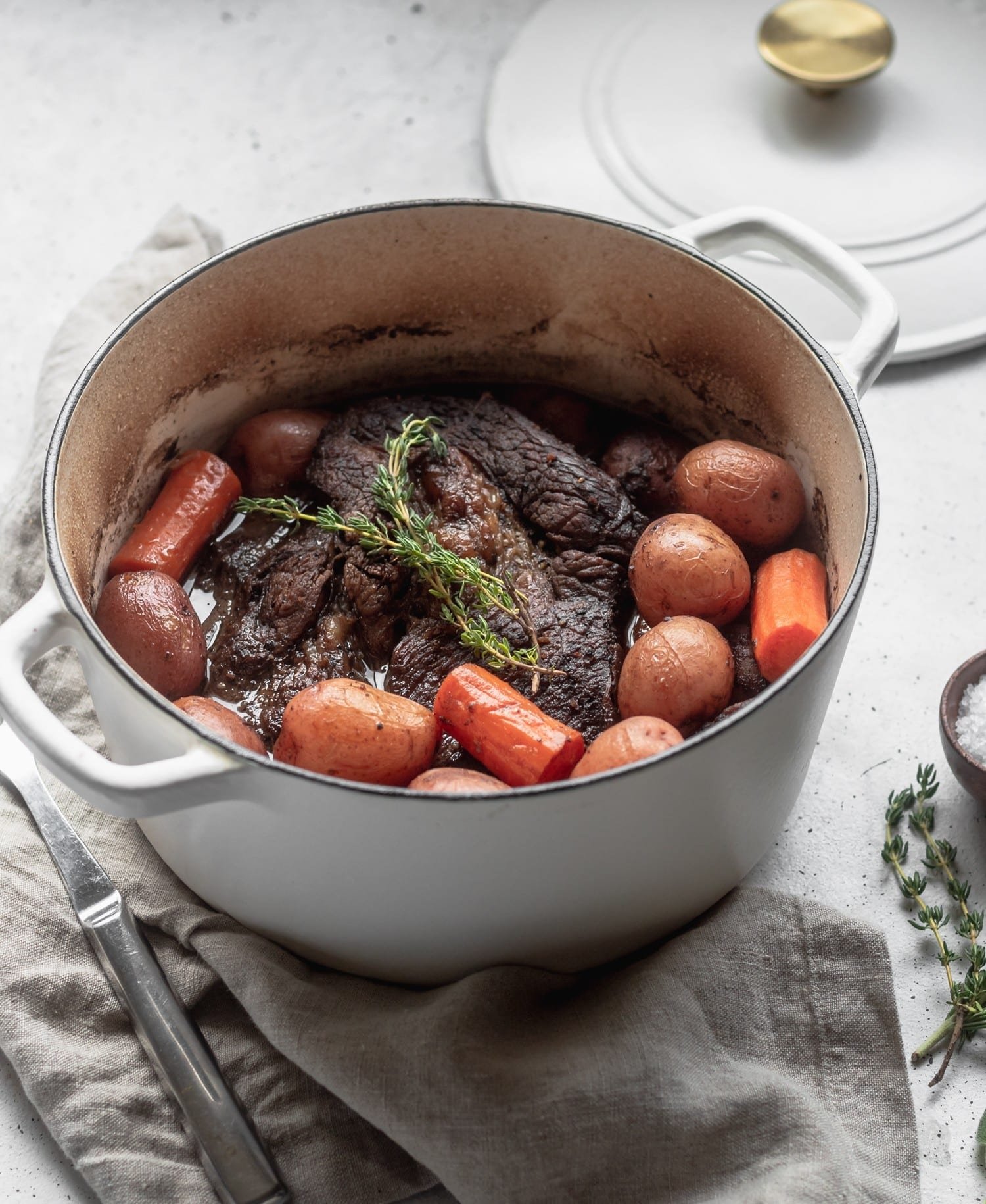 A 45 degree shot of a white pot filled with beef and vegetables on a beige linen with a white background.