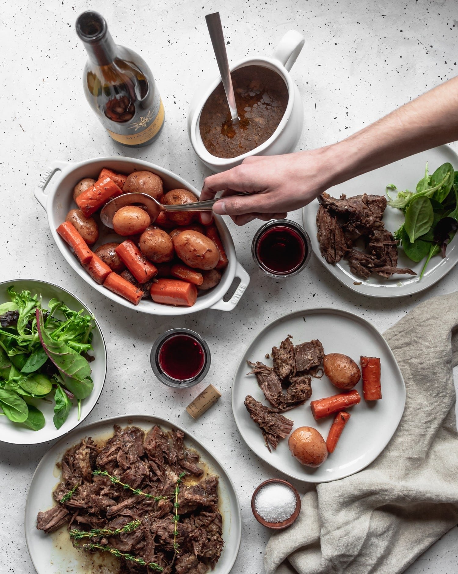 A plate of pot roast surrounded by a plate of potatoes and carrots and a bowl of salad on a grey background. A man's hand is spooning out some potatoes on a plate.