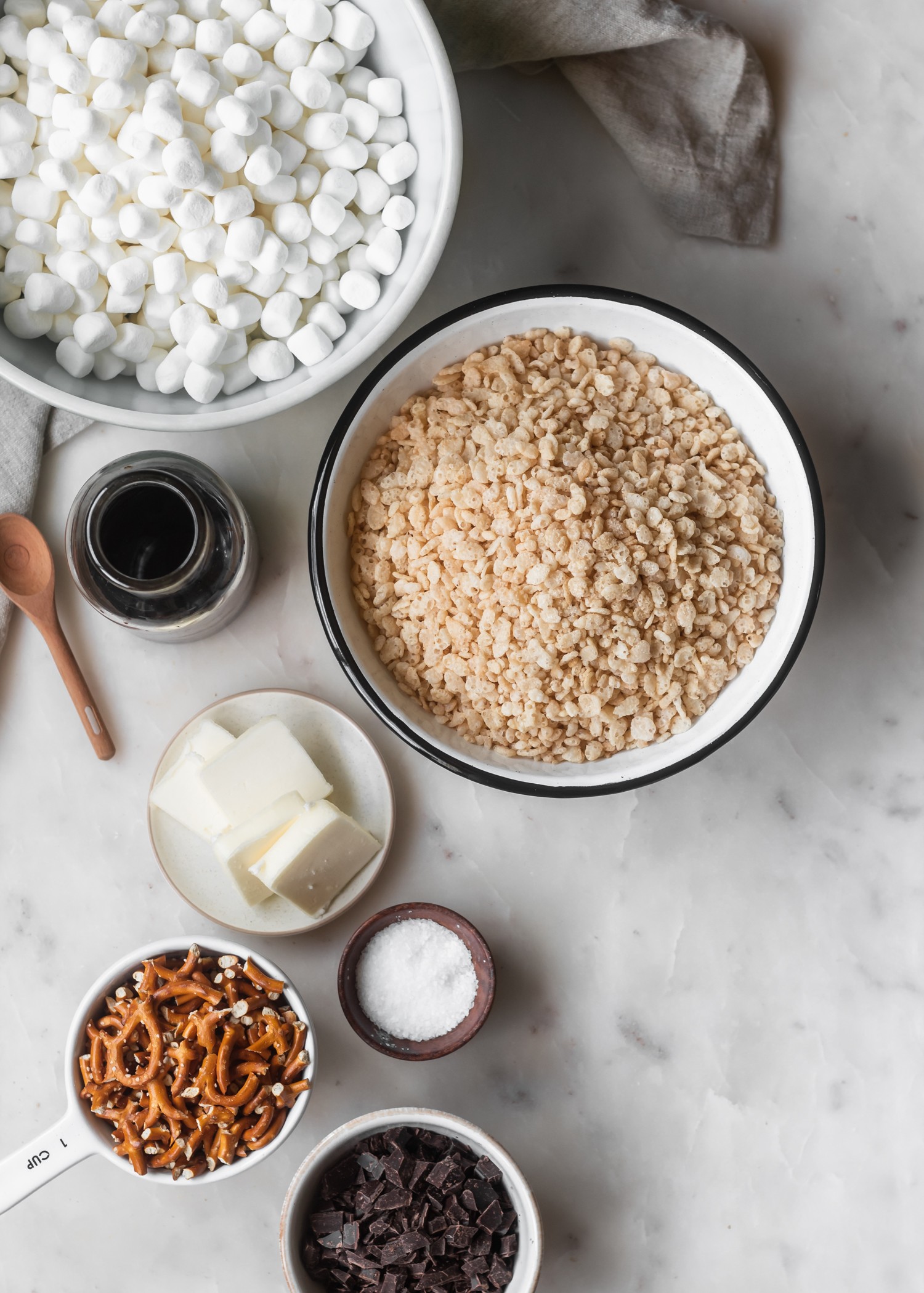 Rice Krispie treat ingredients in white bowls on a white marble background with a beige linen in the corner.